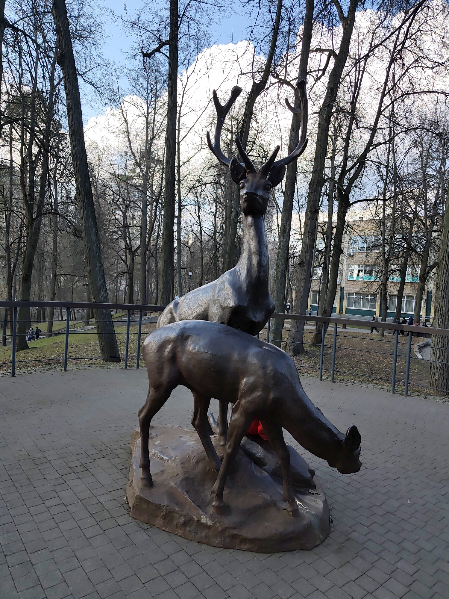 A bronze statue of two deer stands on a brick-paved area within a park in Ramenskoye, Russia. The larger deer stands tall with antlers, while the smaller one is depicted bowing its head. The surrounding area includes trees, a metal fence, and a building in the background. Several people are visible in the distance near the fence. The sky is blue with scattered clouds.