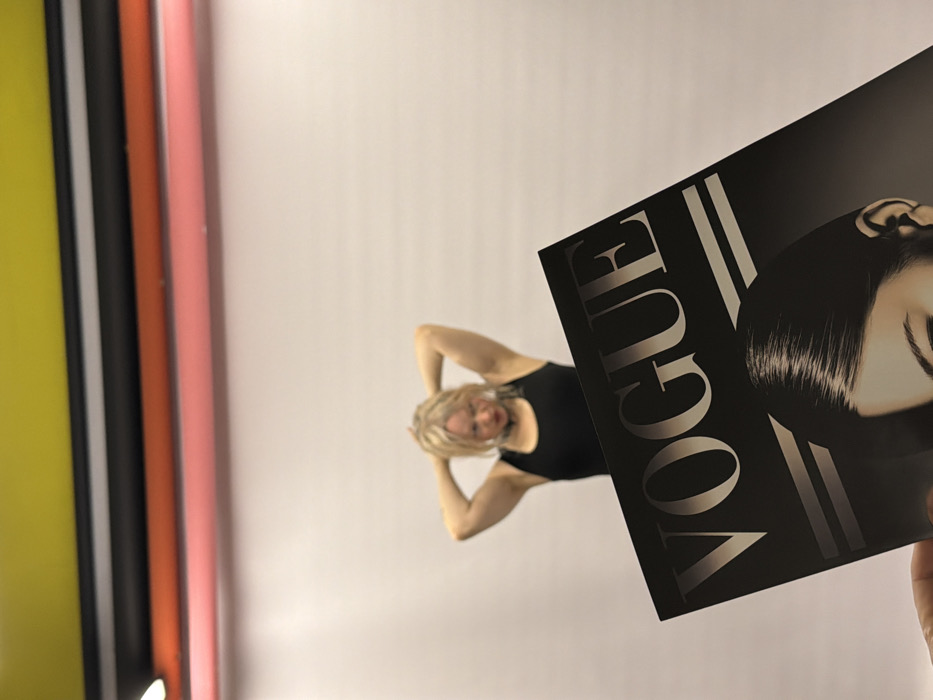 The scene depicts an indoor photography studio located in Saint Petersburg, Russia. In the immediate foreground, a light-skinned hand holds a "VOGUE" magazine at an upward angle, occupying the lower left portion of the frame. The visible cover features the "VOGUE" logo in light lettering on a dark background, above a monochrome image of a woman's forehead, eye, and ear. Behind the magazine, an individual with blonde hair, dressed in a black tank top and a dark choker, stands out of focus. Their hands are raised and clasped behind their head in a posing stance. The background consists of a plain, light-colored studio wall or backdrop. Above the backdrop, multiple horizontal rolls of material are visible, stacked from top to bottom in yellow, black, orange-red, and pink. A studio light fixture is partially visible on the far left. No other subjects or interactions are present.