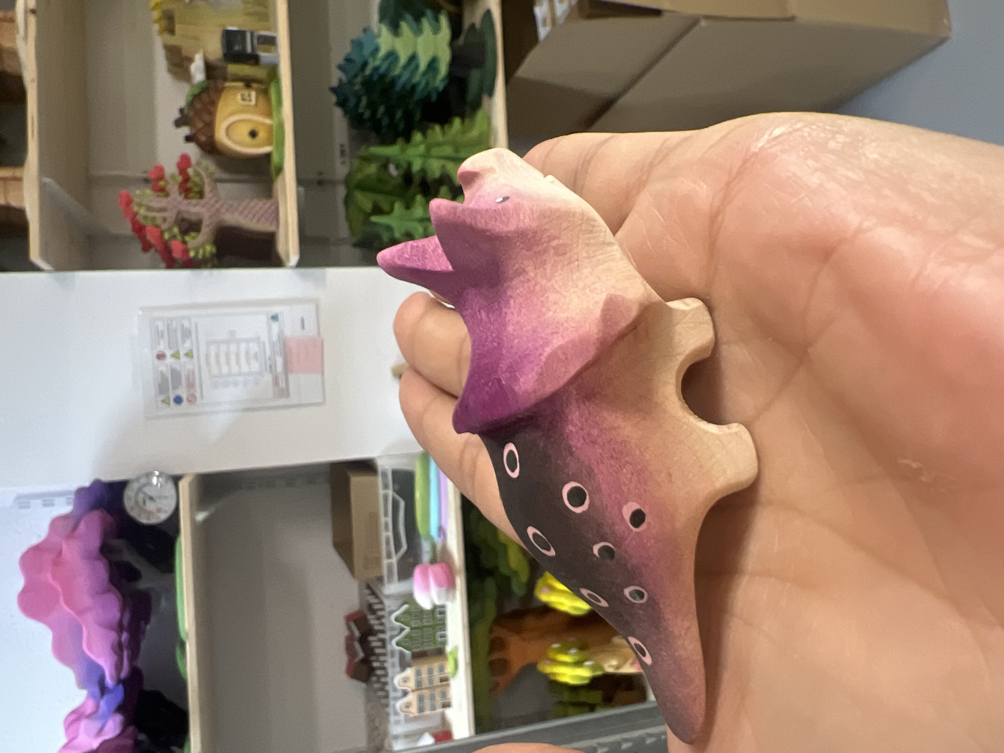 A wooden toy Triceratops dinosaur, painted purple and pink with black polka dots, is held in a person's open palm. The dinosaur has a distinct frill and three horns. In the background, shelves are visible, filled with various other toys and decorative items, including small houses, trees, and abstract cloud-like sculptures. A thermometer is also mounted on one of the shelves. The scene appears to be indoors, possibly a retail store or a playroom, with a neutral background color.