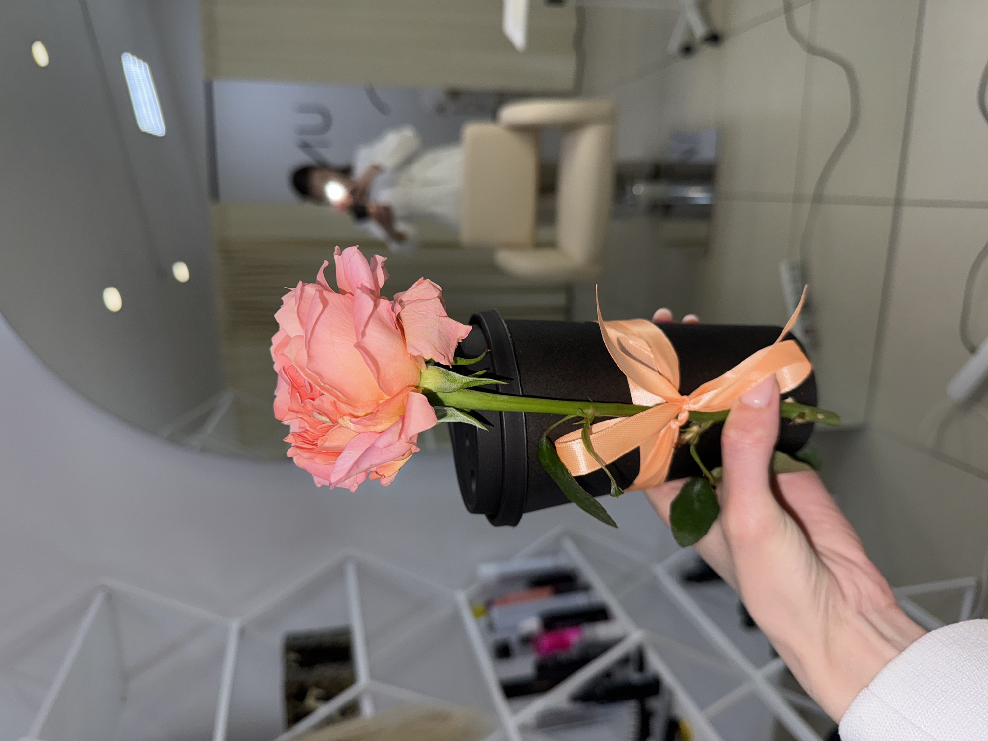 A close-up shot depicts a hand holding a black cylindrical object with a light peach-colored rose emerging from one end, tied with a matching ribbon at the other end. The background reveals a blurred interior scene, possibly a salon or studio, featuring a person seated in a chair and a makeup table with various items. The location is identified as Chaikovsky, Russia.