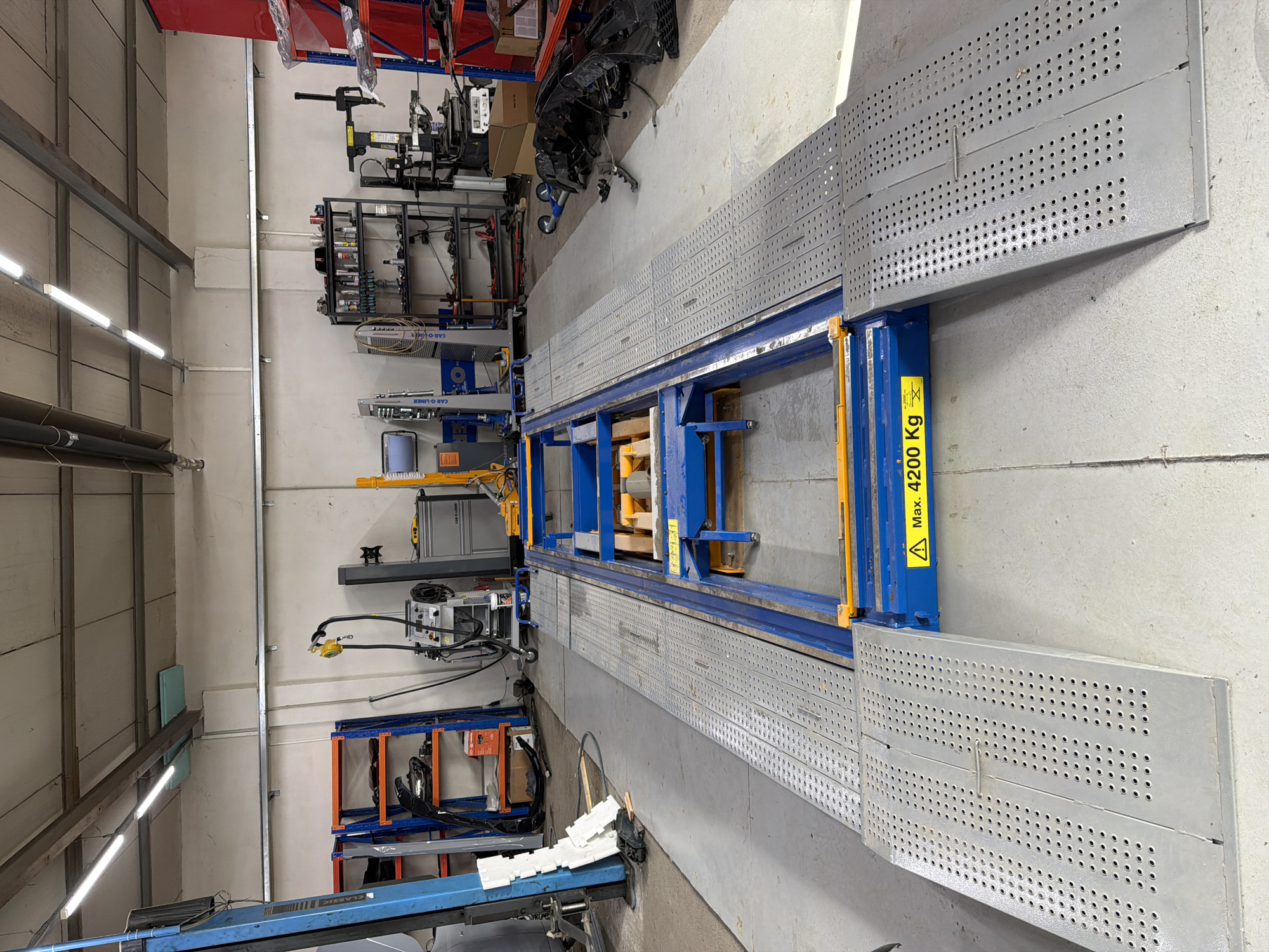 The image displays an interior view of what appears to be an automotive repair or body shop, characterized by its industrial setting and specialized equipment. The central focus is a large, robust, blue and yellow frame straightening or car repair bench. This heavy-duty machinery features long, grey perforated metal platforms or ramps extending from its sides, designed for vehicles to be driven or placed upon. A prominent yellow warning label on the side of the main blue structure reads "Max. 4200 Kg," accompanied by a black exclamation mark inside a yellow triangle, indicating its maximum load capacity.
In the background and around the main equipment, various tools, racks, and automotive components are visible. There are shelves holding a variety of supplies and smaller tools, and what looks like a black car bumper or front fascia component resting on the floor near a blue and red shelving unit. Other specialized machinery, potentially for welding, measuring, or bodywork, can be seen mounted on walls or stands. Text such as "CAR-O-LINER" and "CLASSIC" is visible on some of the blue equipment, likely identifying brand names or models.
The facility has light-colored walls and a concrete floor. Bright fluorescent strip lights are mounted on the ceiling and walls, providing clear and even illumination throughout the space, suggesting it is daytime or that artificial light is the primary source. Large pipes run along one wall, indicating utility infrastructure. The overall impression is one of an organized, professional workshop, well-equipped for vehicle maintenance and repair. No people are present, and no specific activity is taking place; the scene is static, showcasing the operational readiness of the shop.