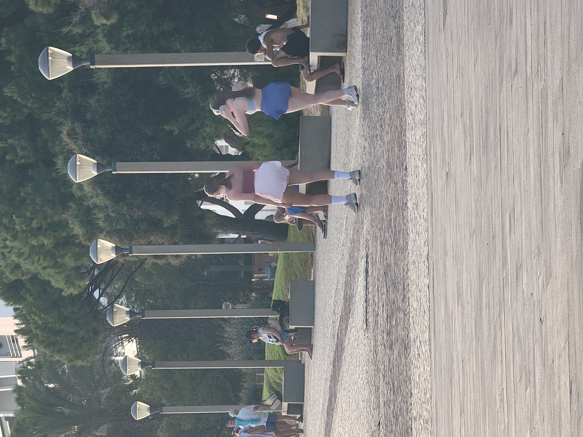 The image depicts a scene on a paved walkway near a body of water, likely the Atlantic Ocean, in Quarteira, Portugal. In the foreground, two young women are walking away from the camera. The woman on the left is wearing a reddish-pink halter top and a short, voluminous white skirt, with white socks and grey sneakers. The woman on the right is wearing a light blue sports bra and a blue skirt, with headphones over her ears and grey sneakers. A man with dark hair is sitting on a grey concrete bench to the right, wearing dark shorts and a white hooded jacket. In the background, several lampposts line the walkway. Further back, a woman in a black hat and sunglasses is sitting on a bench with a dog at her side. Other people are visible in the background, some standing and others sitting on benches. Lush green trees and a white building are visible behind the walkway. The sunlight suggests it is daytime, casting shadows on the paved surface.