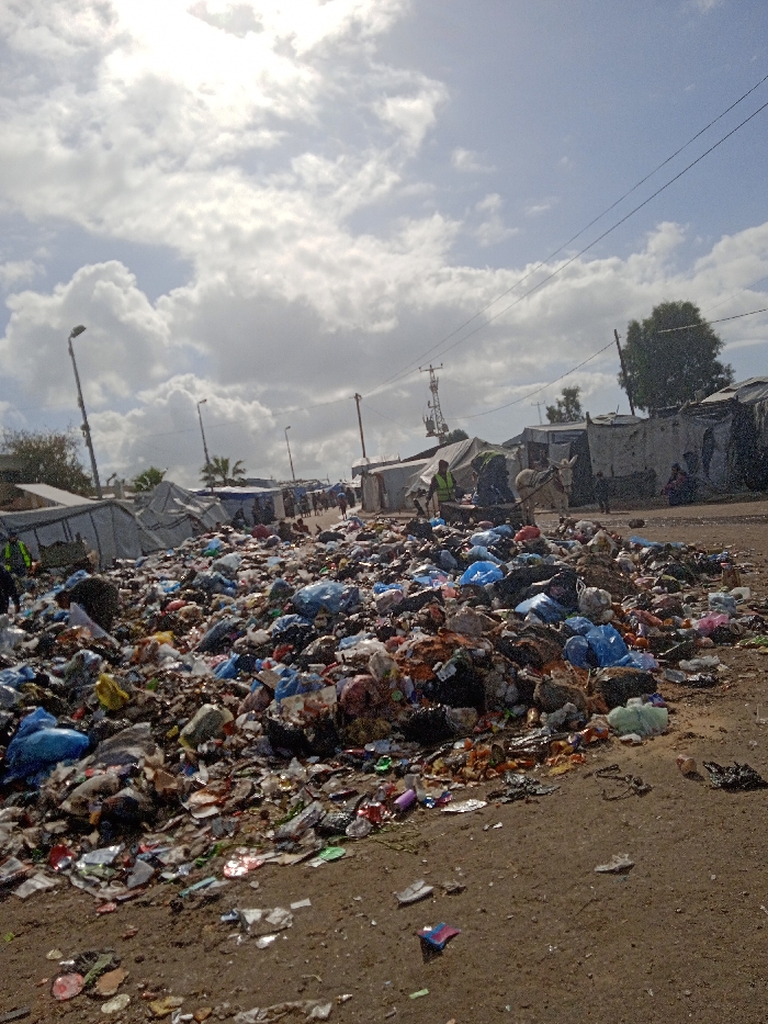 The image captures an outdoor scene in Khan Yunis, Palestinian Territory, dominated by a substantial, unmanaged refuse pile under a partially cloudy, bright sky. A large mound of mixed waste, comprising numerous blue and black plastic bags, assorted packaging, and general debris, occupies the foreground and center, resting on a dirt ground surface that appears wet in areas. In the background, a dirt thoroughfare is flanked by a dense arrangement of rudimentary shelters constructed from tarpaulins, corrugated metal, and other salvaged materials, extending into the distance on both sides. Several individuals are present: one person wearing a reflective vest is partially visible near the garbage pile on the left. Further back on the right, a white donkey pulls a two-wheeled cart laden with materials, guided by another person in a reflective vest. A third individual is seated on the ground near the shelters on the far right. Multiple tall streetlights and utility poles with overhead power lines are visible along the thoroughfare, indicating infrastructure within the crowded settlement environment.