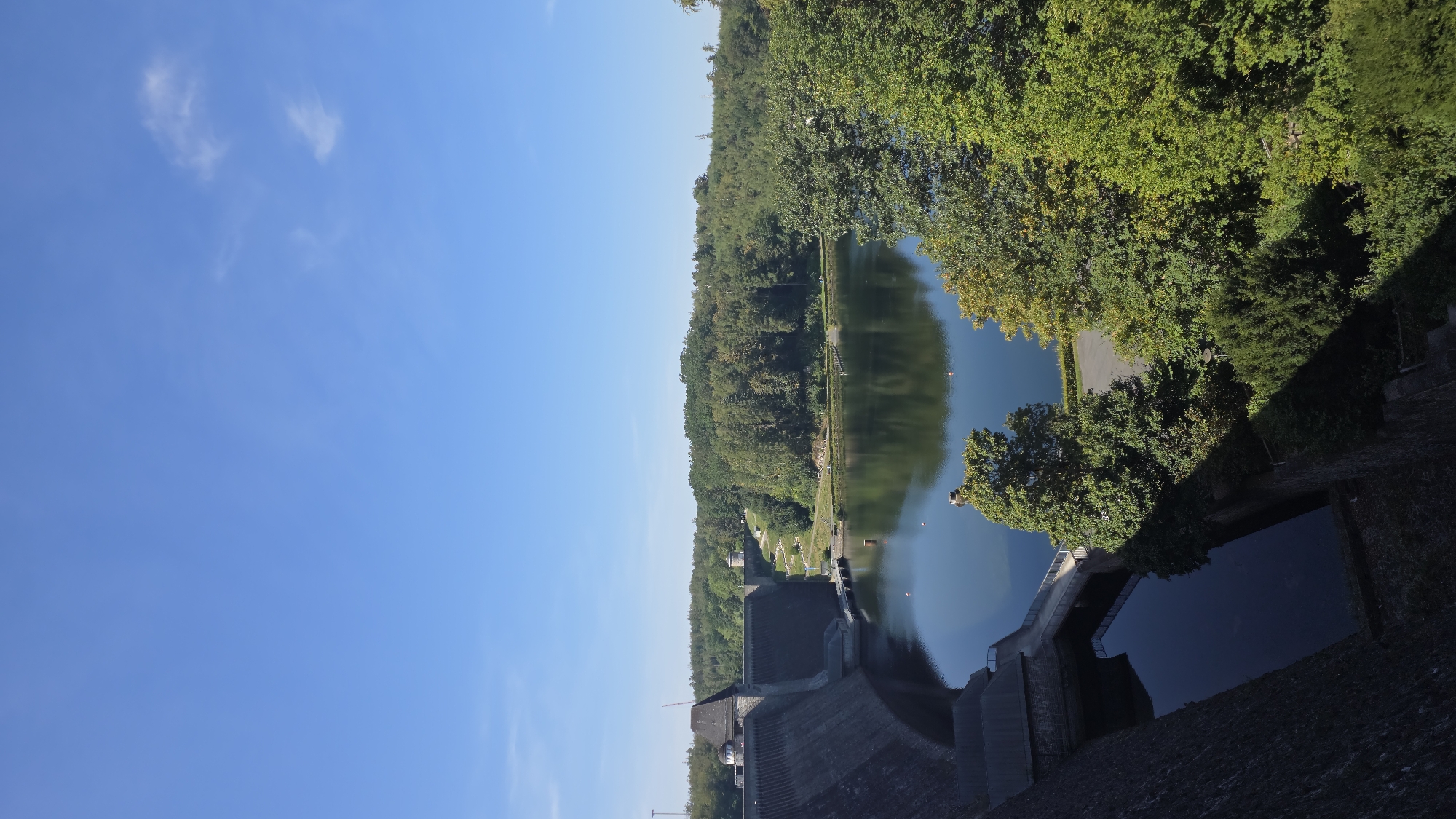 The image captures a scenic view of the Möhnesee dam in Möhnesee, Germany. The dam, constructed of stone and concrete, dominates the left side of the frame, with its curved structure holding back a large body of water. The reservoir behind the dam is calm and reflects the surrounding lush green trees and the clear blue sky.
The dam itself features several architectural elements, including towers and walkways. A pathway is visible leading away from the dam towards a hill covered in dense forest. The trees are a mix of deciduous and coniferous species, with vibrant green foliage indicating it is likely summertime. The sky is a brilliant blue with a few wispy white clouds scattered across it.
The foreground shows the rough, textured surface of the dam, with some vegetation growing on its lower sections. The water in front of the dam appears dark and deep. The overall impression is one of natural beauty combined with impressive engineering. There are no people visible in the image.