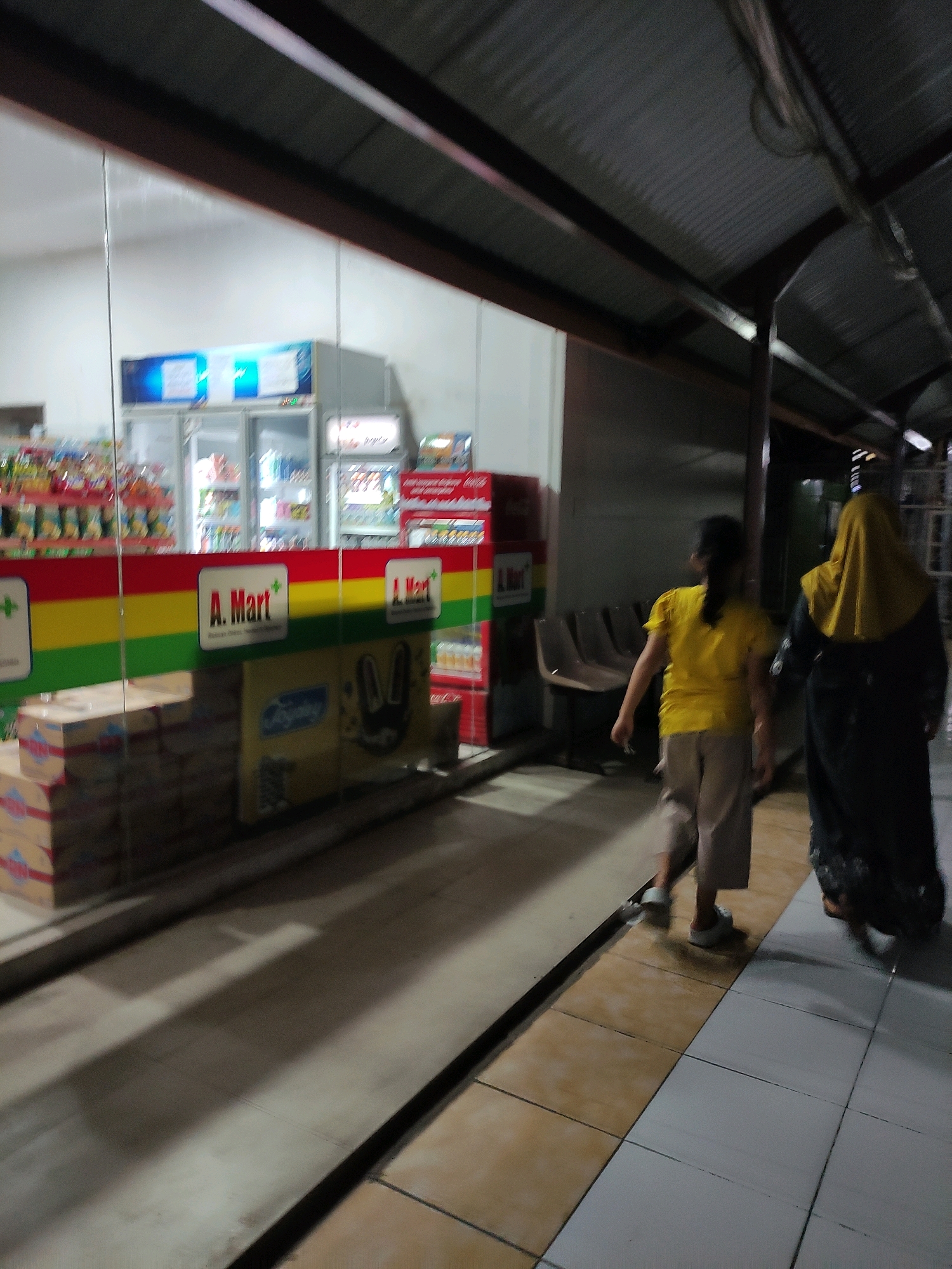 The image depicts the exterior of an A. Mart convenience store in Bulukumba, Indonesia. The store has a glass storefront revealing refrigerators and shelves stocked with products. A prominent A. Mart logo is visible on a red, yellow, and green striped banner. Boxes are stacked on the ground outside the store. Two individuals are walking away from the store along a tiled pathway. One person wears a yellow shirt and light-colored pants, while the other is dressed in a dark outfit with a yellow headscarf. The overhead structure suggests a covered walkway or an outdoor market setting. The lighting indicates it is either dusk or nighttime.