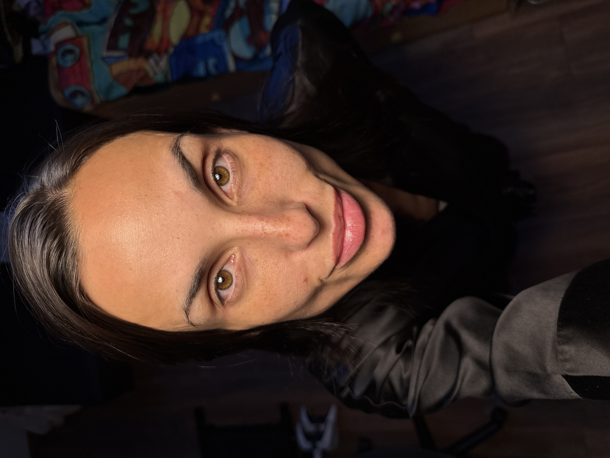 The image is a close-up, high-angle shot of a woman's face. She has brown hair and brown eyes, with visible freckles on her skin. She is wearing a black satin robe or jacket. The background is dark, with some indistinct objects visible, including what appears to be patterned fabric on the right side. The image does not provide any information about the location in Pleven, Bulgaria.
