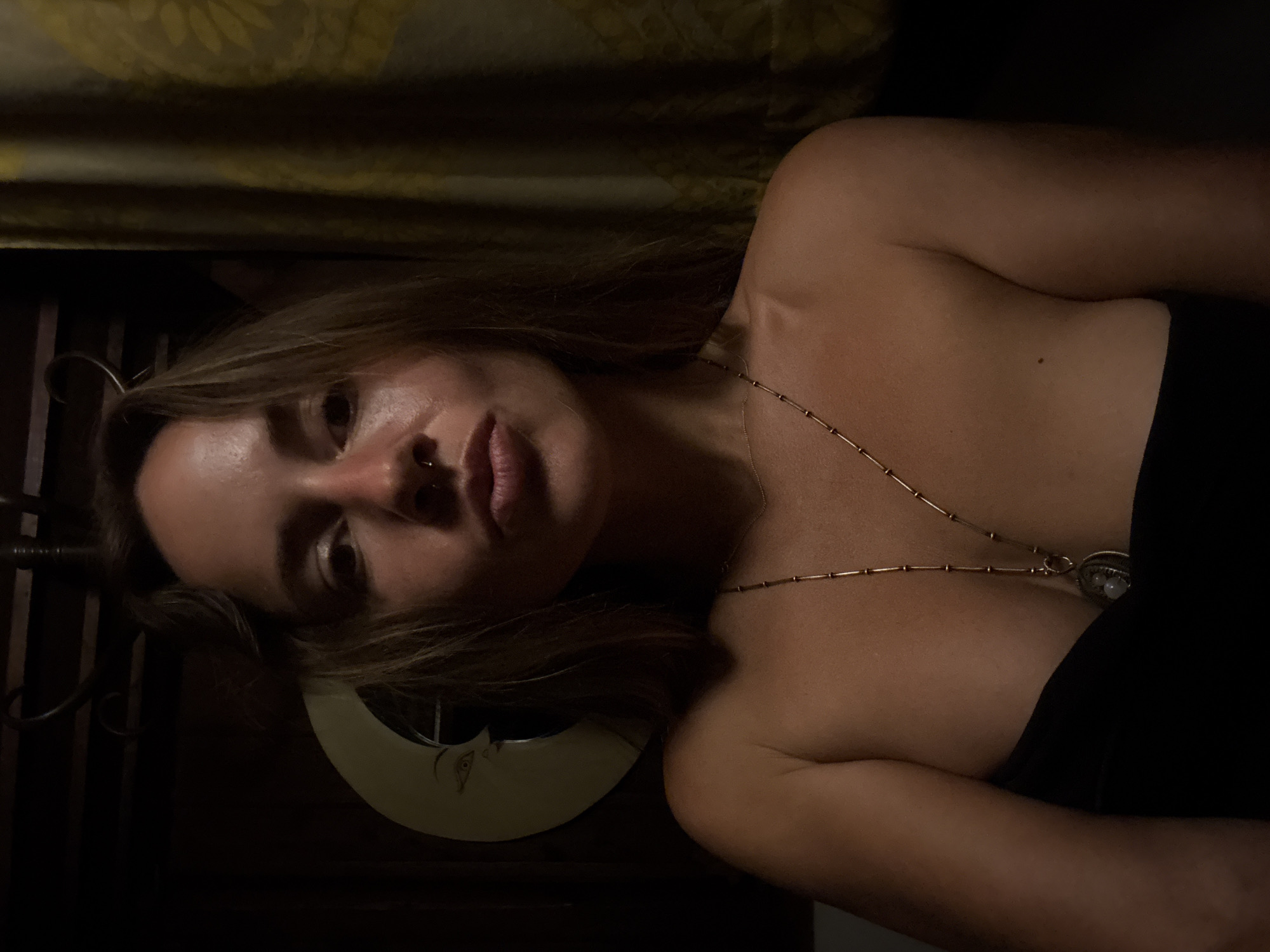 A woman is pictured from the shoulders up, looking directly at the camera with her lips pursed. She has long, brown hair and a nose ring. She is wearing a strapless black top and a gold necklace with a pendant. The background is dark and dimly lit, with a yellow curtain visible on the right and a circular decoration resembling a moon on the left. The overall lighting is warm and atmospheric.
