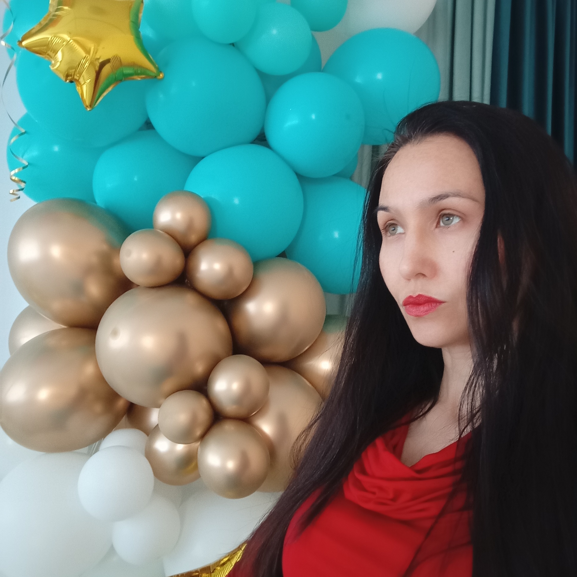 A woman stands in front of a balloon arrangement, possibly in a room in Izhevsk, Russia. The balloon arrangement is comprised of gold, teal, and white balloons, including a gold star-shaped balloon at the top. The woman has dark hair and is wearing a red garment. She is looking off to the side. The background includes a white wall and a portion of what appears to be a curtain.