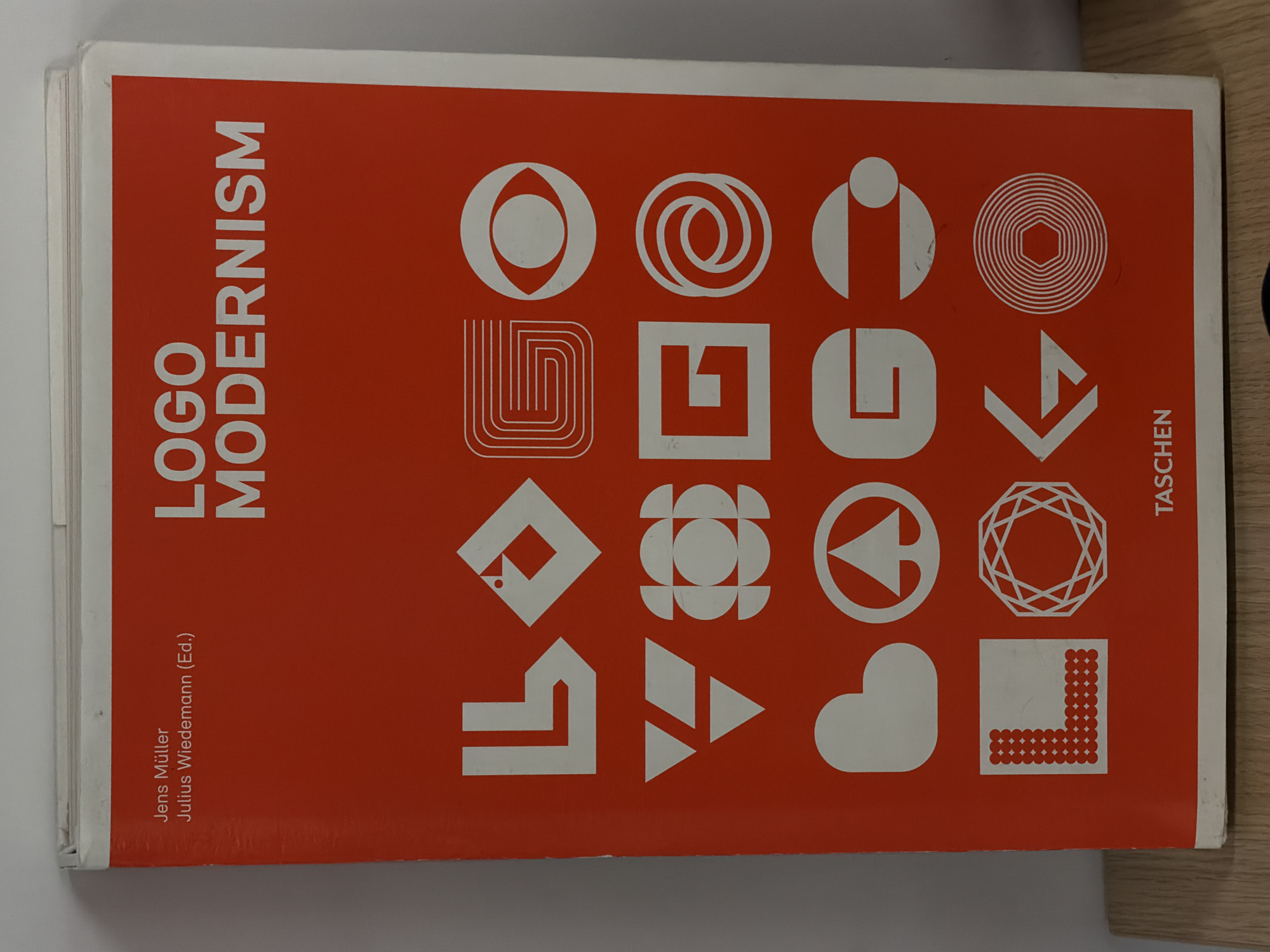 The image shows a book titled "Logo Modernism" by Jens Müller and Julius Wiedemann (Ed.). The book has a red cover with a collection of white logos arranged in a grid. The word "TASCHEN" is printed vertically on the right side of the book. The background is a mix of white and wooden surfaces. The location in Yerevan, Armenia, is not determinable from the image.