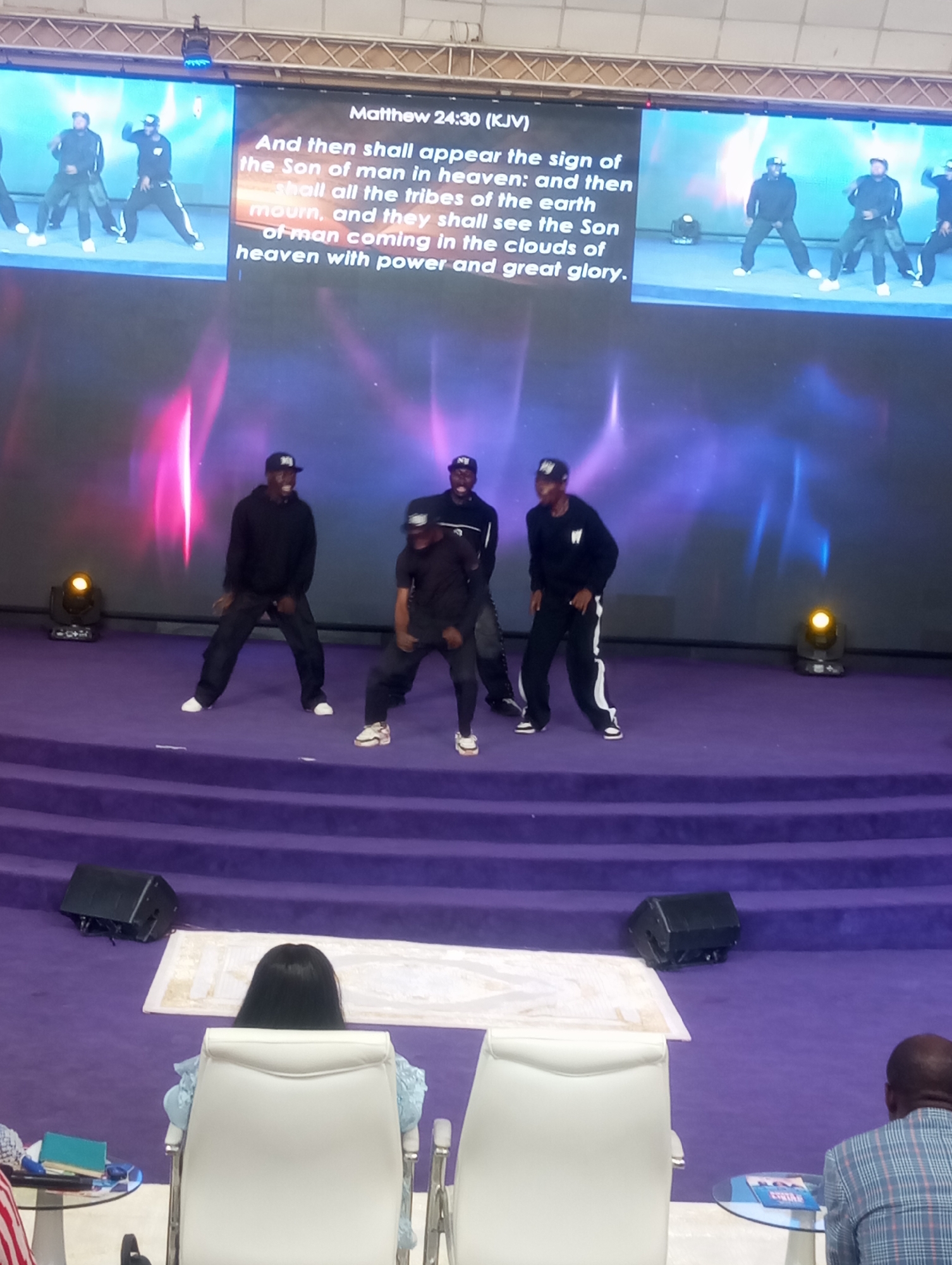 The image shows a presentation on a stage in what appears to be a church or auditorium. On a large screen behind the stage, a video or image is playing, and biblical text from Matthew 24:30 (KJV) is displayed. Below the screen, four men are dancing on the stage. They are wearing black clothing and caps. The stage is covered in purple carpet. In front of the stage, there are rows of seats with a few people sitting and watching. Some lighting equipment is visible on and around the stage. The image was taken in Gura Top, Nigeria.