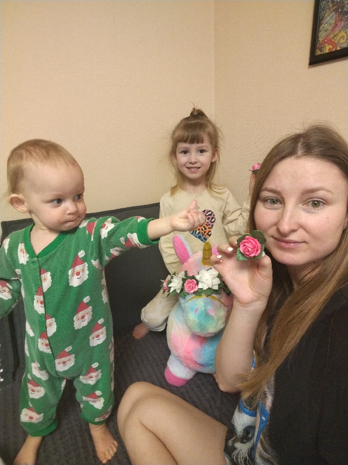 The image captures three individuals – a woman and two young children – in an indoor setting, likely a home, with a plain light-colored wall in the background. They appear to be sitting or standing on a dark-colored sofa or couch.
On the right side of the frame, a woman with long, straight brown hair and green-hazel eyes is visible from the chest up. She has fair skin and is wearing a dark-colored top, possibly black. She is smiling faintly and looking directly at the camera. Her right hand is raised near her face, holding a small, intricate decorative pink rose with green leaves. Another similar pink rose is tucked into her hair on the right side of her head.
In the middle background, a young girl with light brown hair, styled with bangs and a small bun on top, is standing. She is also smiling broadly at the camera. She is wearing a long-sleeved light-colored shirt featuring a large, colorful heart design made of various animal print patterns. She is barefoot.
To the left, in the foreground, a baby or toddler with light brown hair is standing, wearing a green full-body sleepsuit patterned with multiple white Santa Claus faces and red hats. The baby is looking towards the left with a slight pout or smirk and has its right arm extended, seemingly pointing or reaching out of the frame.
Between the young girl and the woman, a plush toy unicorn is visible. It is multi-colored (pink, blue, purple, yellow tie-dye) and has a golden horn. A decorative flower crown, similar in style to the rose held by the woman, adorns the unicorn's head, featuring small pink and white roses with green leaves.
A framed artwork or picture can be seen hanging on the wall in the upper right corner of the image. The overall impression is a casual, warm family moment, possibly a selfie given the woman's direct gaze and proximity to the camera. The Santa-patterned sleepsuit on the toddler suggests the photo might have been taken around the Christmas season.