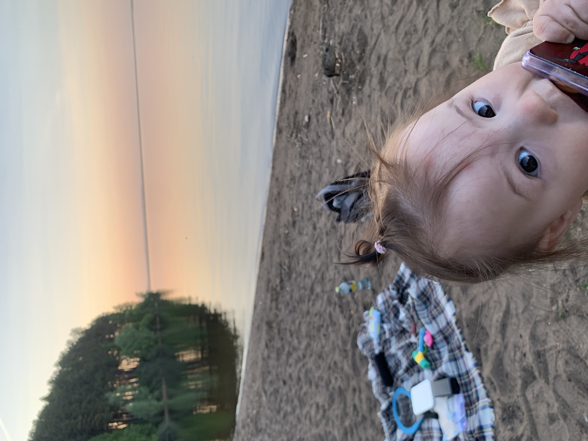 A close-up, rotated, and slightly blurry eye-level shot displays a baby with light brown hair and dark eyes, seemingly chewing on a smartphone. The child is situated on a sandy area, possibly a beach, with a plaid blanket spread out nearby, scattered with toys and a water bottle. In the background, a body of water reflects a sunset, hinting at the edge of a forest. The location is presumed to be a beach in Chaikovsky, Russia, though this cannot be confirmed from the image itself. The primary subject is the baby, and the secondary subjects are the blanket and shoreline objects. The action is the child chewing on a phone, and the environment is outdoors, specifically a beach setting.