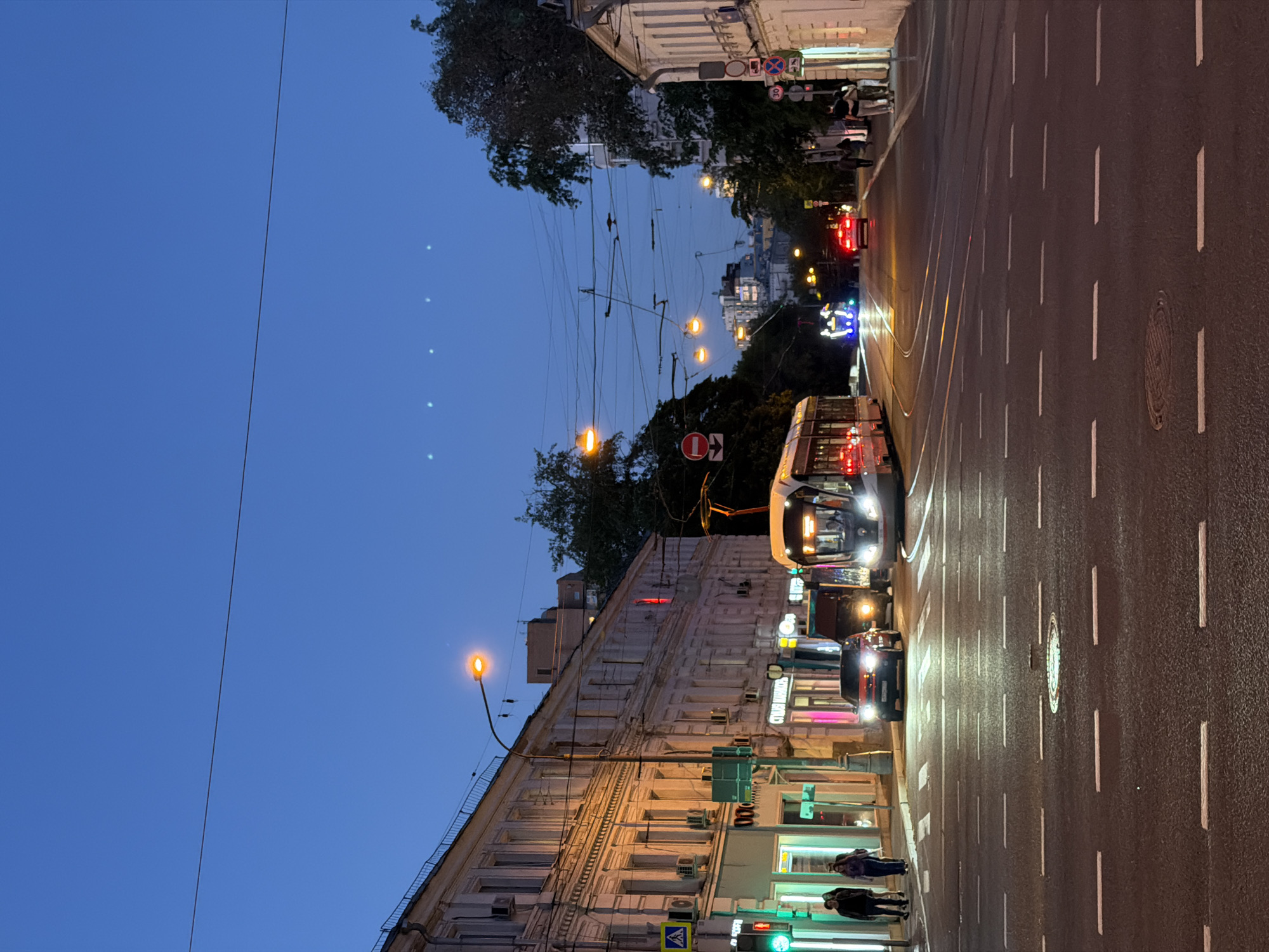 The image shows a street scene in Moscow, Russia, at dusk. A tram and several cars are on the street. There are people on the sidewalk. Buildings line the street. There are street lights. The sky is a deep blue.