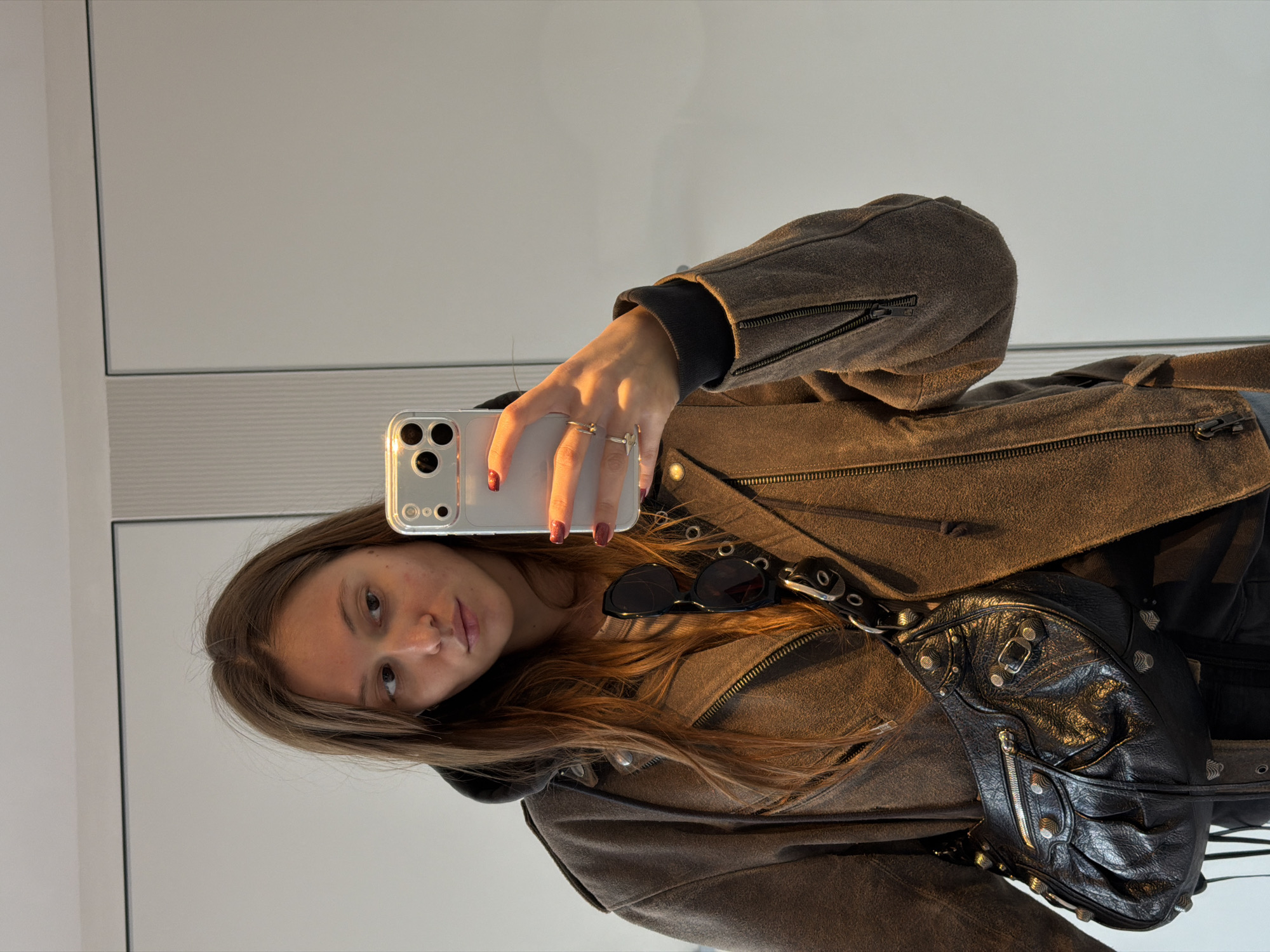 A woman is taking a selfie in a mirror. She is wearing a brown suede jacket over a black hoodie, with a black studded purse slung over her shoulder. Sunglasses rest on the jacket. The background appears to be a plain white wall, likely indoors. There is no indication of the location being Moscow, Russia.
