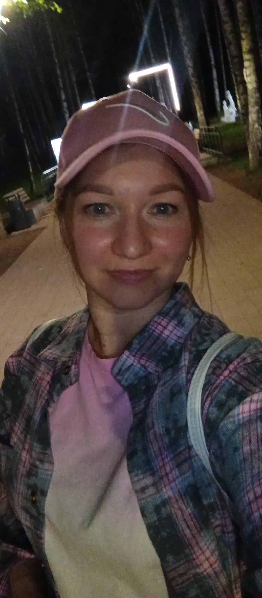 A woman is pictured in an outdoor setting in an unknown city. She is wearing a pink baseball cap, a pink t-shirt, and a gray plaid shirt. Behind her are trees and a walkway, and a lighted structure. The lighting suggests it is night.