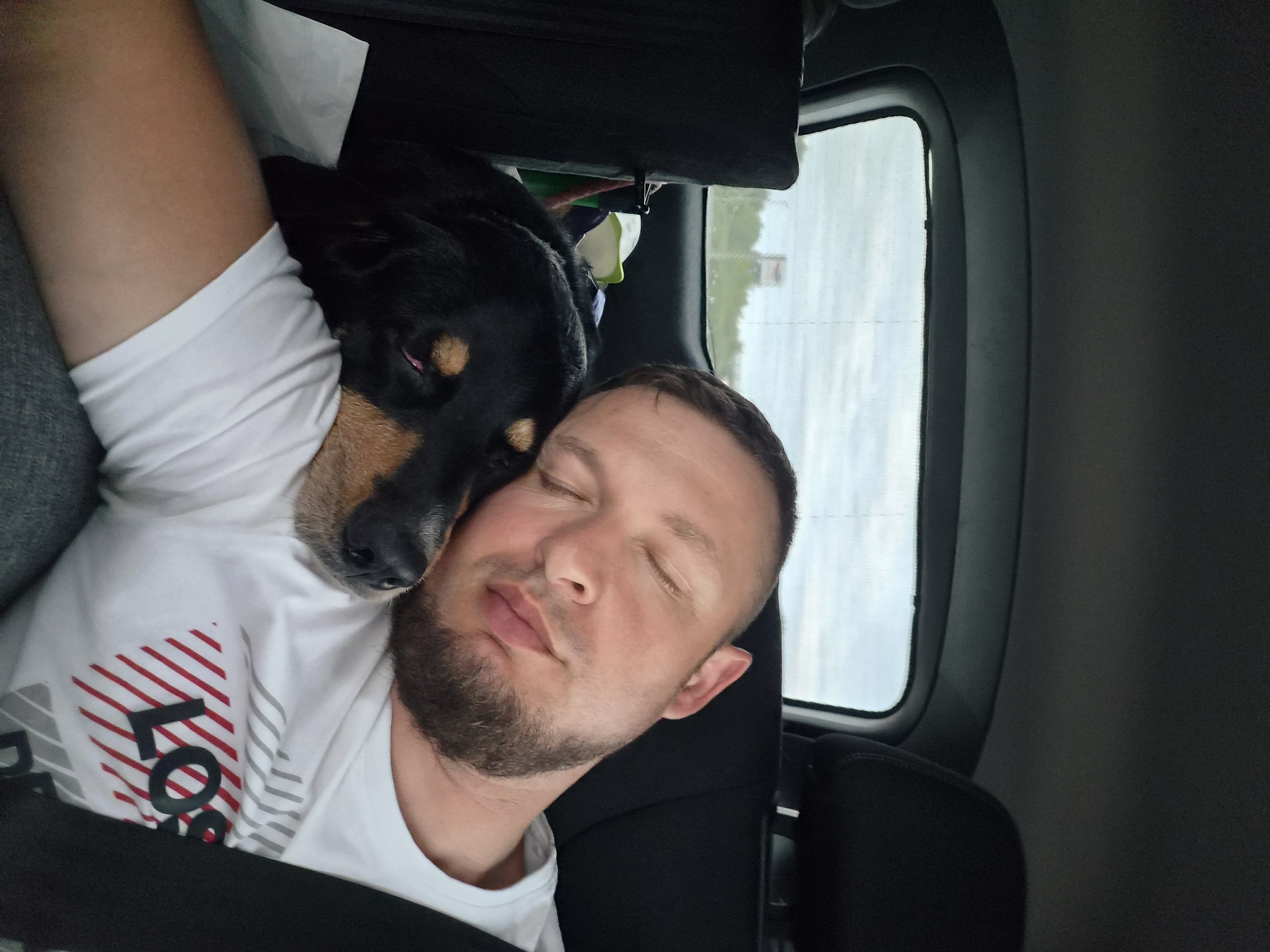 A man and a dog are asleep in the back seat of a vehicle. The man is on the right, with his eyes closed and his head resting on his arm. His beard is short and dark. He is wearing a white t-shirt with red and gray diagonal stripes and the partial word "LOS" visible. The dog is on the left, a dark-colored breed with tan markings around its muzzle and eyes. The dog's head is resting on the man's chest and shoulder, and its eyes are closed. The background shows the interior of a car, with the rear window revealing a cloudy sky and trees and a sign in the distance, suggesting a rural or suburban roadside location. The general setting appears to be the city of Groß Santersleben, Germany.