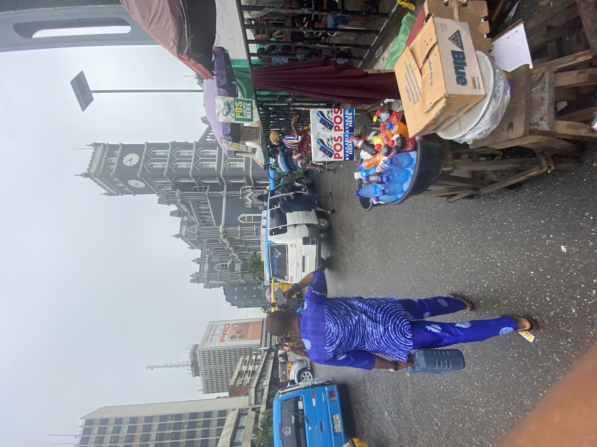 The image shows a street scene in an urban area. A man wearing a blue patterned outfit is standing on the street. He is holding a gray container in his left hand and facing left. In the background, there is a large, ornate clock tower building, and several other buildings of varying heights. There are vehicles on the road, including a blue minivan and a white van. Street vendors are visible with their stalls selling items such as bottled drinks and packaged goods. Signage is also visible, including one promoting POS (Point of Sale) availability. The sky is overcast, suggesting potentially wet or humid weather. The street is wet. There are several people standing around the road and a truck parked in front of the church.