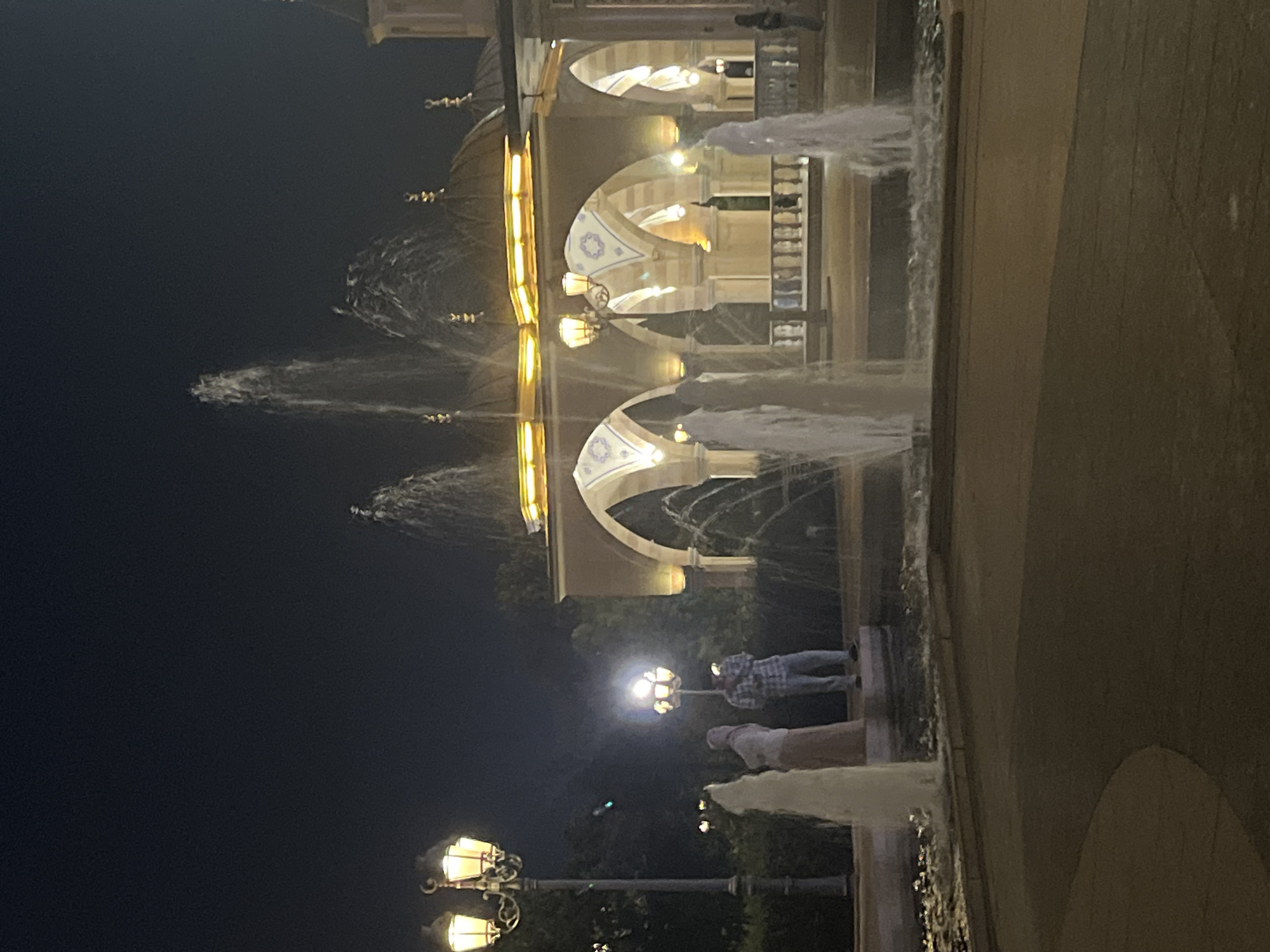 The image captures a nighttime scene at a fountain in Grozny, Russia, likely in front of a building with arched architecture, possibly a mosque or a government building. A tall lamppost with multiple glowing bulbs stands to the left, illuminating the pathway and a portion of the fountain. In the foreground, water jets from a fountain spray upwards, creating a misty effect.
Two individuals are visible near the lamppost on the left. One person, wearing a headscarf and a long dress, stands to the left, facing away from the camera. Another person, dressed in a plaid shirt and jeans, stands to the right of the first individual, appearing to hold a phone and looking towards the building.
The building in the background features a series of arches supported by columns. Golden lights illuminate the building's facade, highlighting its architectural details, including domes and decorative elements on the roof. The overall ambiance is that of a well-lit public space at night, with the water feature adding a dynamic element to the static architecture. The ground appears to be paved.