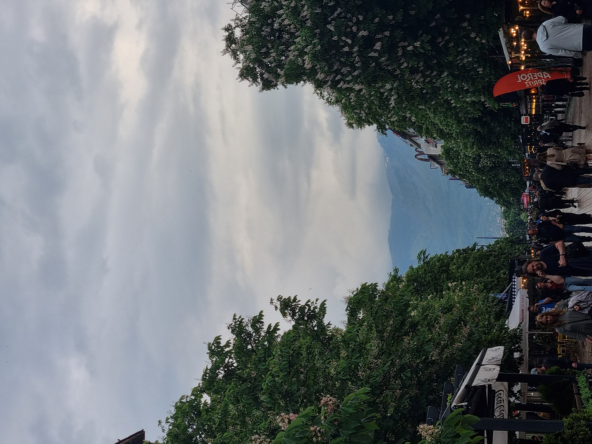This image depicts a vibrant outdoor scene on a tree-lined street in Sofia, Bulgaria, during what appears to be late afternoon or early evening under an overcast sky. The street is bustling with numerous people strolling, socializing, and enjoying the outdoor setting.
The scene features a wide promenade flanked by tall, leafy trees, possibly horse chestnuts, adorned with white blossoms. In the background, a large mountain range is visible beneath the cloudy sky.
On the right side of the street, there are various outdoor establishments, indicated by tables, seating, and overhead string lights that are illuminated, suggesting the time of day or a festive ambiance. Many individuals are present, some walking, others standing or seated. A prominent orange banner or umbrella, which reads "APEROL SPRITZ" (mirrored), is visible. Further down, a white canopy clearly displays the text "WELCOME TO SOFIA," confirming the location context. Below this, a sign with "Coca-Cola" branding is also discernible.
The overall mood is lively and social, with people casually dressed and engaged in leisure activities. The diffused light from the overcast sky creates soft shadows across the scene.