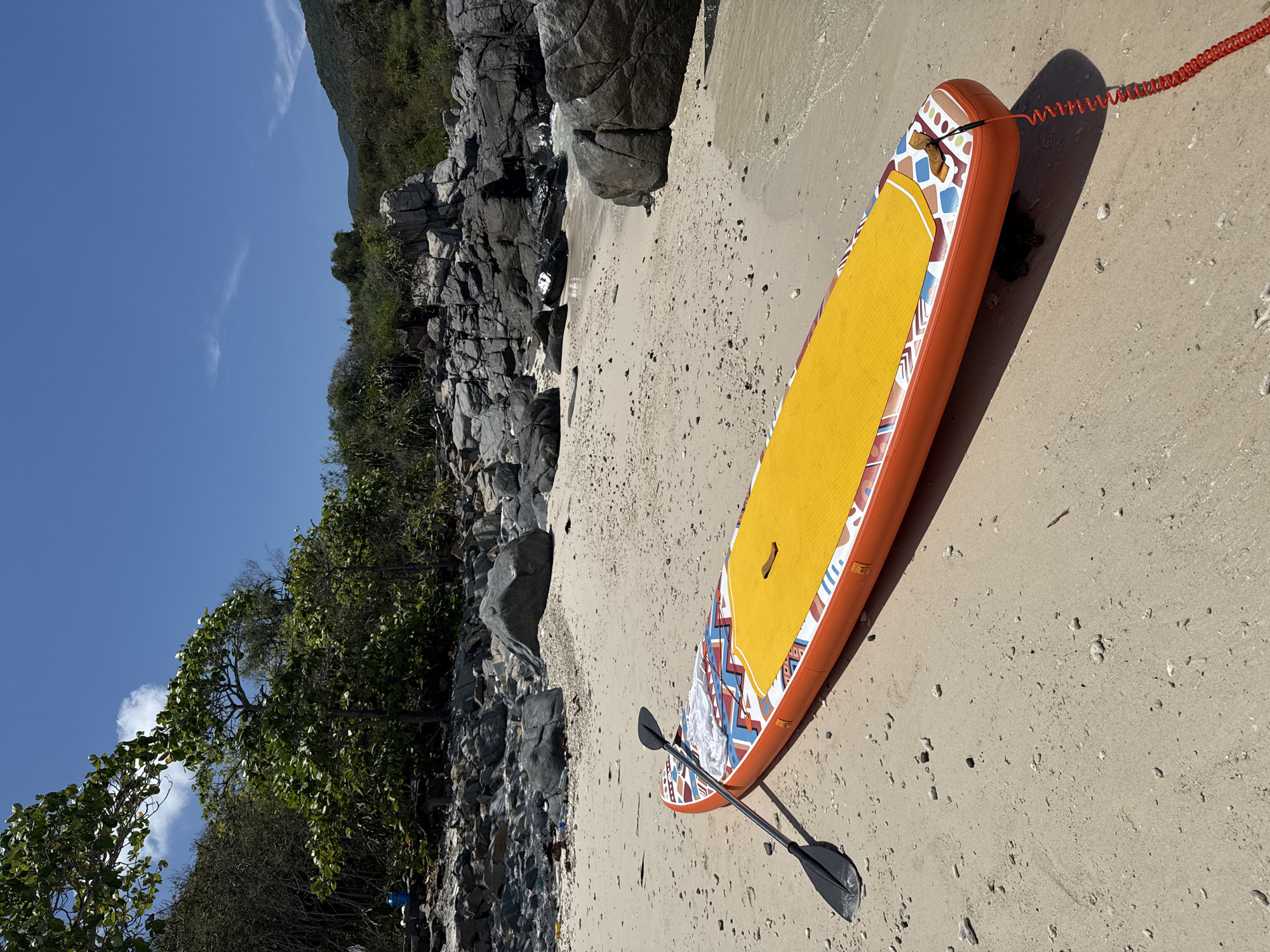 A stand-up paddleboard (SUP) rests on a sandy beach adjacent to a rocky outcropping and vegetation under a clear, blue sky. The paddleboard is orange with a yellow standing pad and patterned trim. A black paddle lies beside the board, and a red coiled leash is attached to the tail. The beach is light-colored sand with scattered small rocks and debris. The location is Nha Trang Ward, Vietnam, evident from the tropical flora, rock formations, and beach characteristics. No people are visible in the image.