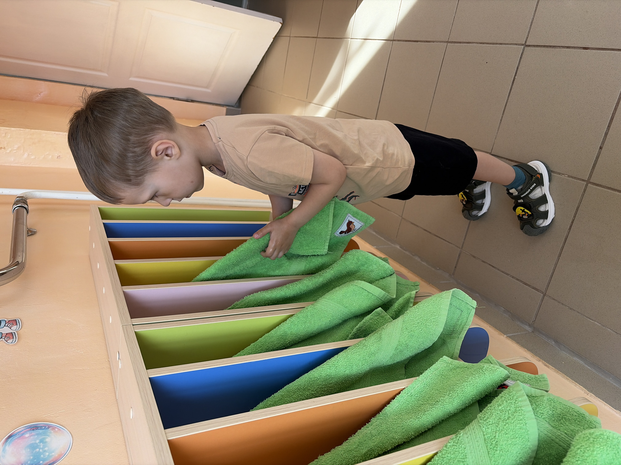 A young child is standing in front of a cabinet with multiple colorful shelves, each containing a folded green towel. The child is wearing a beige t-shirt, black shorts, and sandals. The shelves are orange, yellow, pink, green, and blue. The floor is tiled. The image was taken in Chaikovsky, Russia.