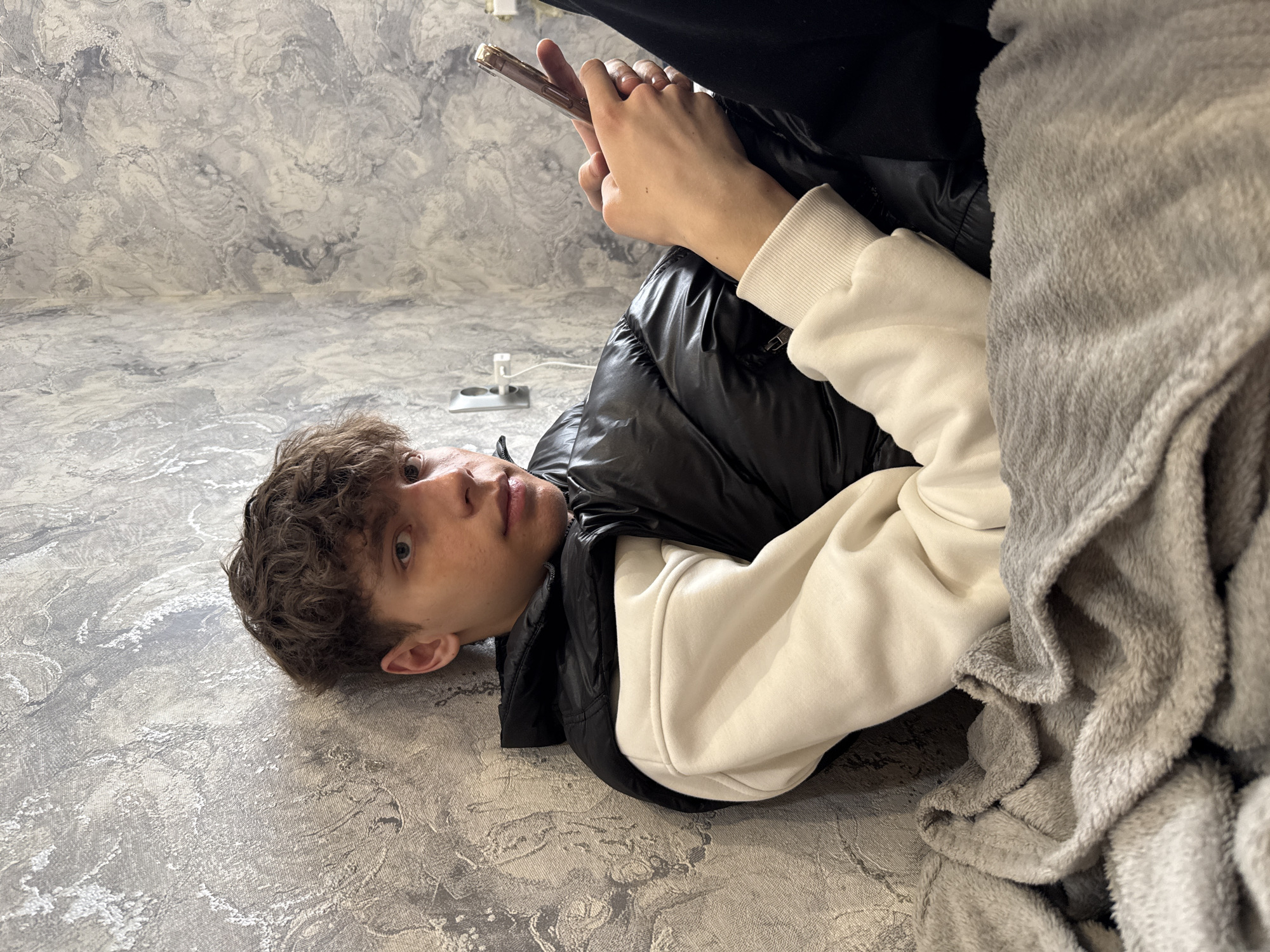 A young man is lying on a textured gray floor, potentially made of concrete or tile. He is wearing a black puffer vest over a light-colored sweatshirt. A gray blanket partially covers his body. He is holding a smartphone in his hands. The background is a gray wall with a mottled, stone-like pattern. An electrical outlet is visible on the floor near the wall. The photo does not contain sufficient information to confirm the specific location as Korobovo, Russia.