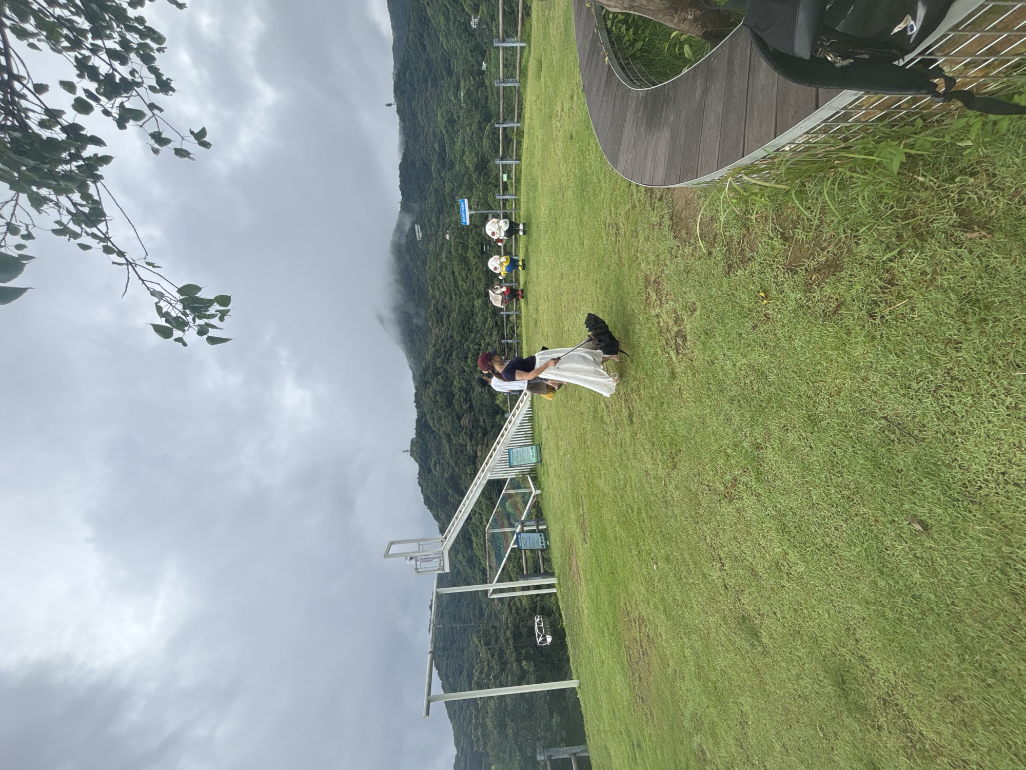 The image depicts a scenic outdoor area with rolling green hills and a cloudy sky. In the foreground, a woman is walking a black dog on a leash across a grassy field. To her left, a white swing set and a slide with a white railing are visible. In the middle ground, several colorful, cartoonish figures are standing on the grass, and a wooden fence encloses the area. In the background, lush green mountains are shrouded in mist, with a few buildings scattered on their slopes. The scene appears to be a park or recreational area, possibly a tourist attraction given the decorative figures. The location is not identified as Guangzhou City, China.