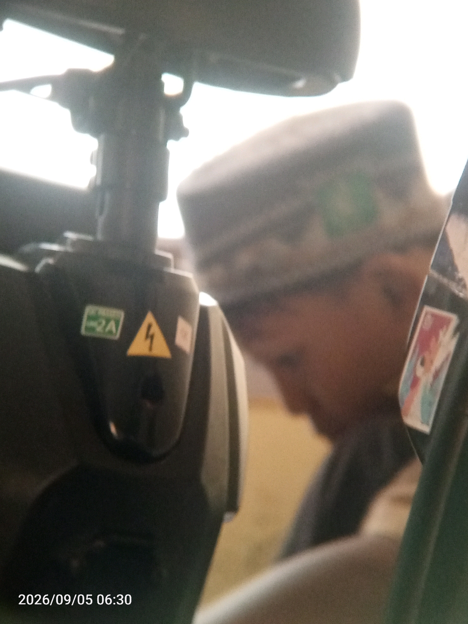 The image is an outdoor close-up shot taken in Randegansari, Indonesia, on September 5, 2026, at 06:30. The focus is on the upper torso and head of a person wearing a patterned cap, blurred and facing to the right. To the left, part of a dark-colored mechanical device is visible, featuring two stickers: one green with "2A" and another yellow with a lightning bolt symbol. A brightly lit sky dominates the background. The overall lighting is bright and diffused, indicating early morning.