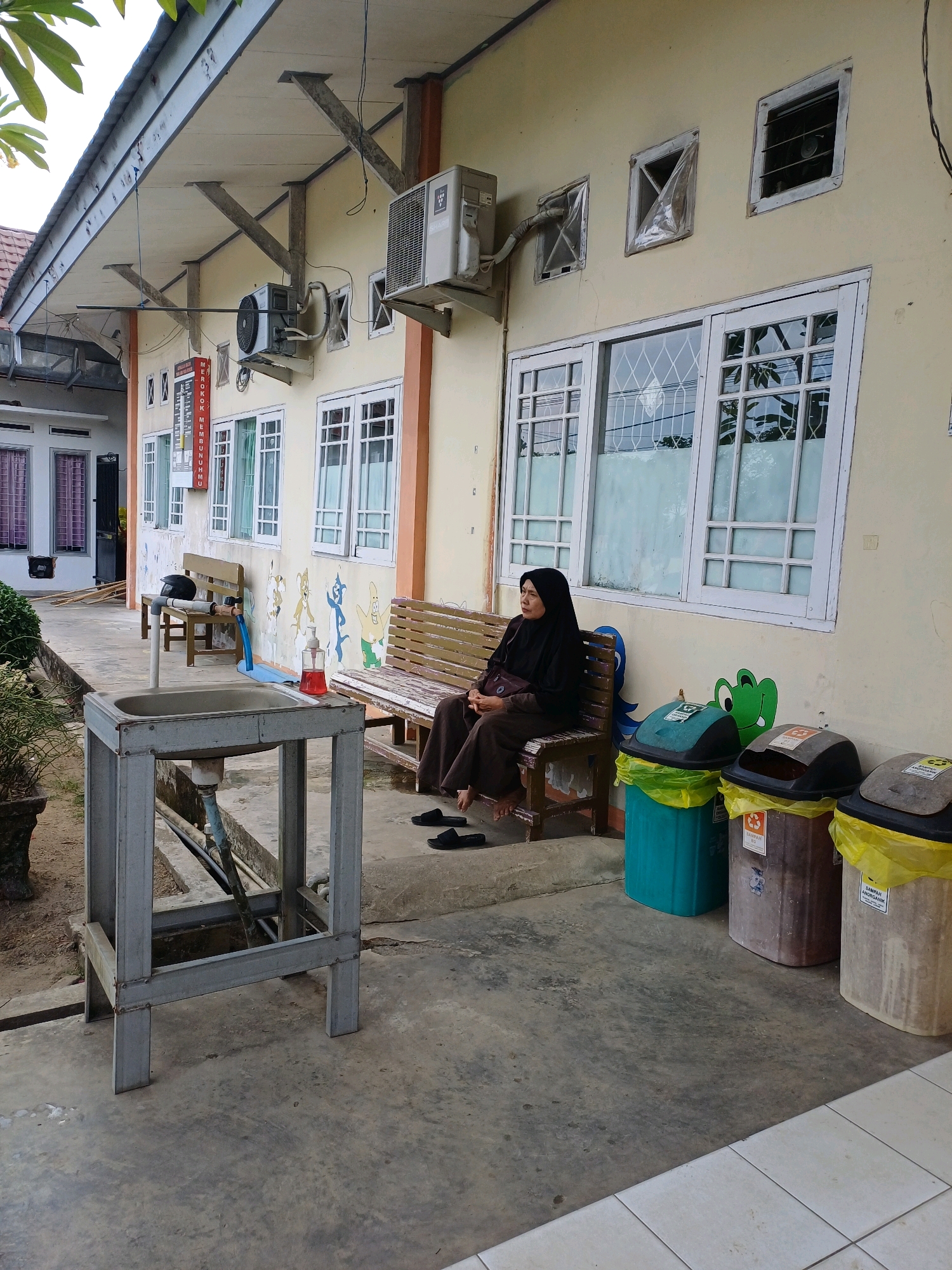 A woman wearing a black hijab and brown clothing sits on a wooden bench outdoors. She is looking to her left. A metal sink with plumbing is in the foreground. Behind the bench, there are three trash cans, two with yellow liners. The scene is set against the exterior wall of a building with several windows. The ground is concrete.