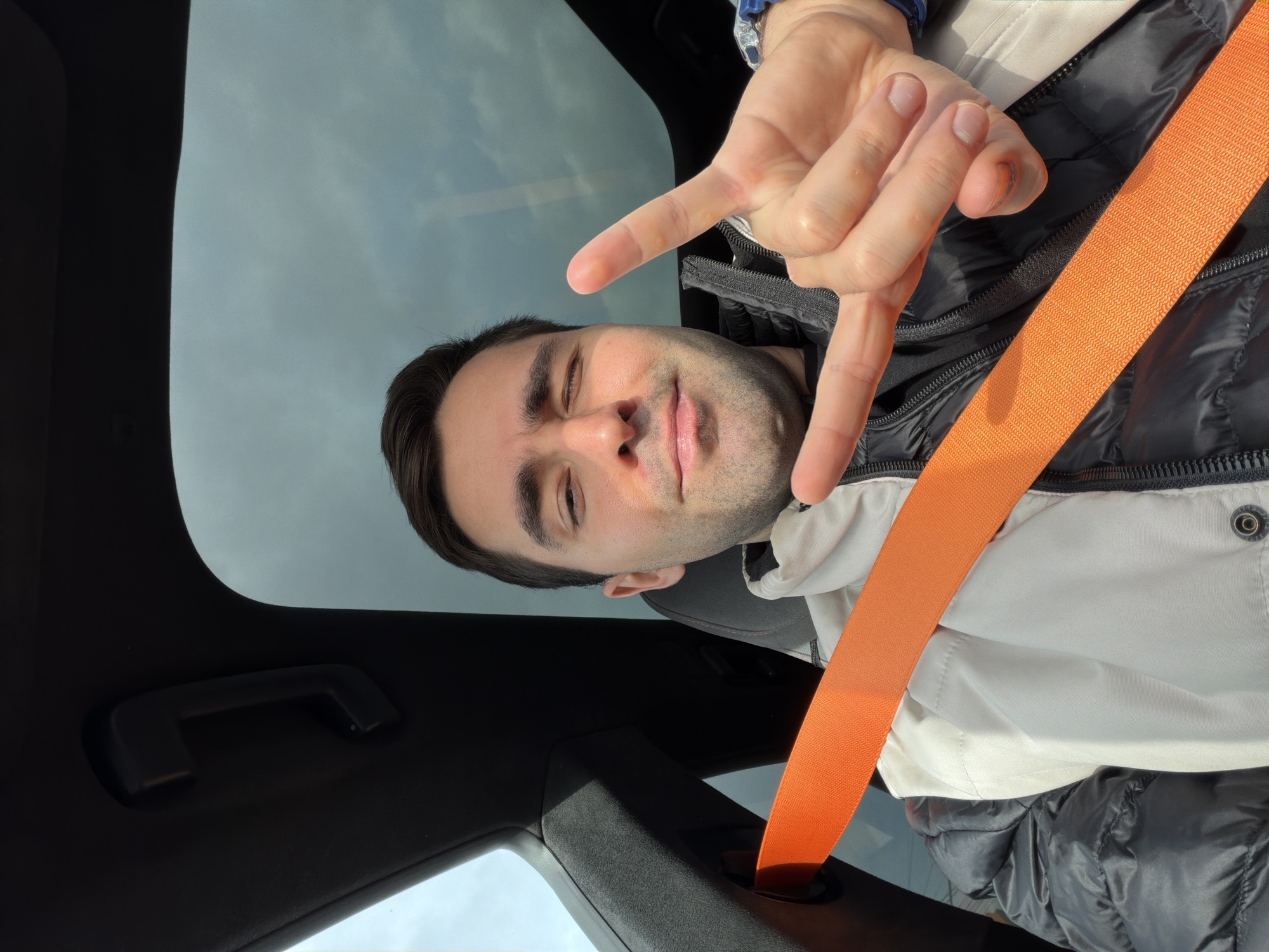 The image, rotated approximately 90 degrees counter-clockwise, captures a man taking a selfie inside a car in Krasnoyarsk, Russia, during the daytime. The man, appearing to be in his 20s or 30s, has short dark hair and light facial stubble. He looks directly at the camera with a slight smile, his eyes partially closed or squinting, possibly from bright light. He wears a dark puffer jacket over a lighter top, and a prominent orange seatbelt crosses diagonally from his right shoulder to his left hip. His right hand is raised, forming a "rock on" gesture with his index and pinky fingers extended. A silver-toned watch is visible on his right wrist.
The background primarily shows an overcast or partly cloudy sky, visible through what appears to be a car's sunroof or windshield. Portions of the car's dark interior are visible, including a black grab handle on the headliner in the upper left, and dark upholstery or dashboard elements at the bottom. The lighting suggests a bright, yet possibly cloudy, day. The overall mood is casual and cheerful. No visible text is present in the image.
