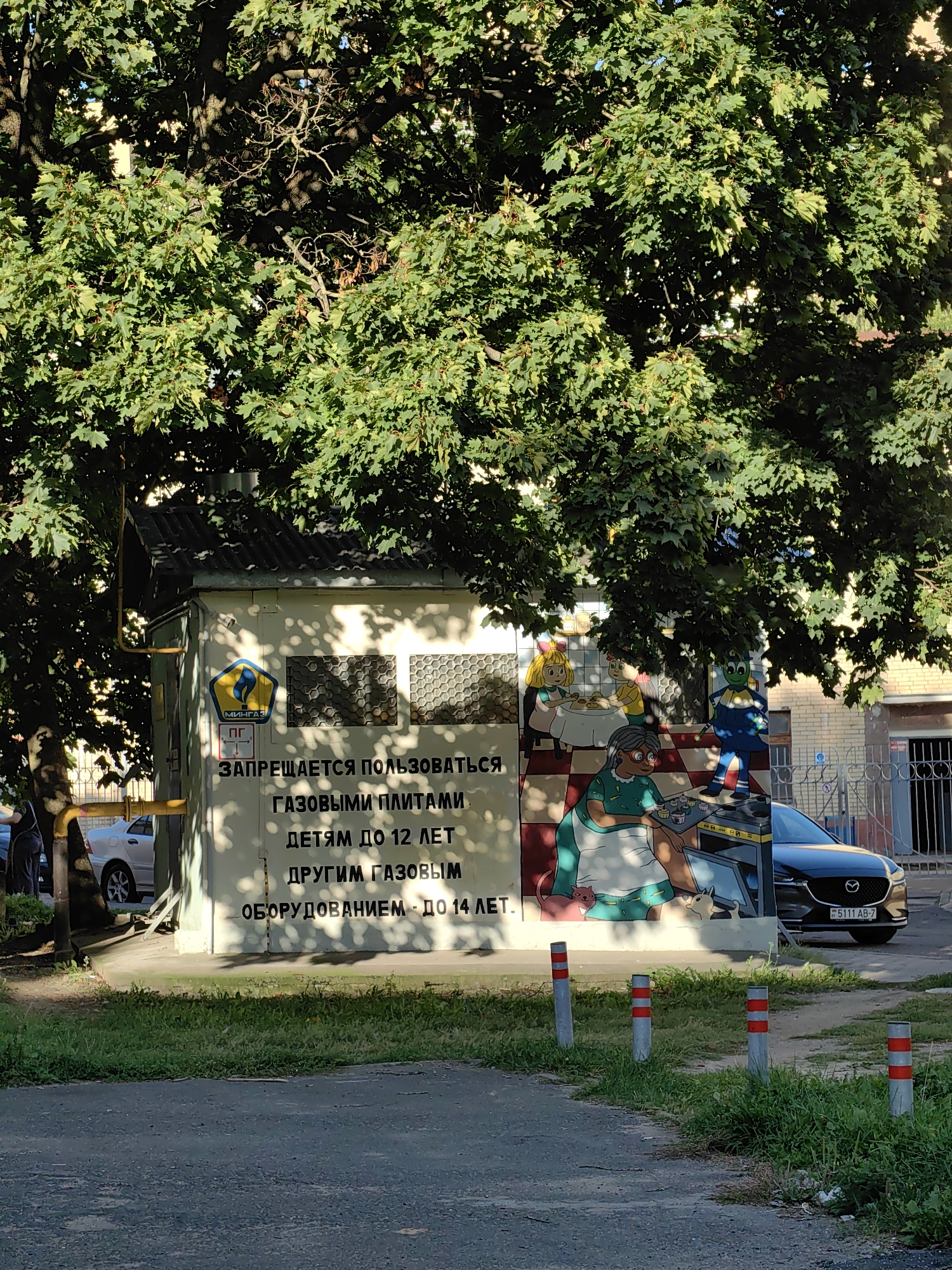 This is a daylight exterior, medium-shot photograph of a gas distribution point in Minsk, Belarus. The structure is a small, rectangular, cream-colored building with a corrugated metal roof. The building is adorned with a mural depicting cartoon characters engaged in domestic activities, possibly related to cooking. In the foreground, a tree with green foliage partially obscures the building. The ground in front of the building is a mixture of asphalt and grass, with several bollards lining the path. A dark-colored car with a Belarusian license plate is parked to the right of the building, and a silver car is parked to the left. There is also a yellow gas pipe running along the exterior of the building and a white fence in the background. The mural contains text in Russian, including a warning about using gas stoves.