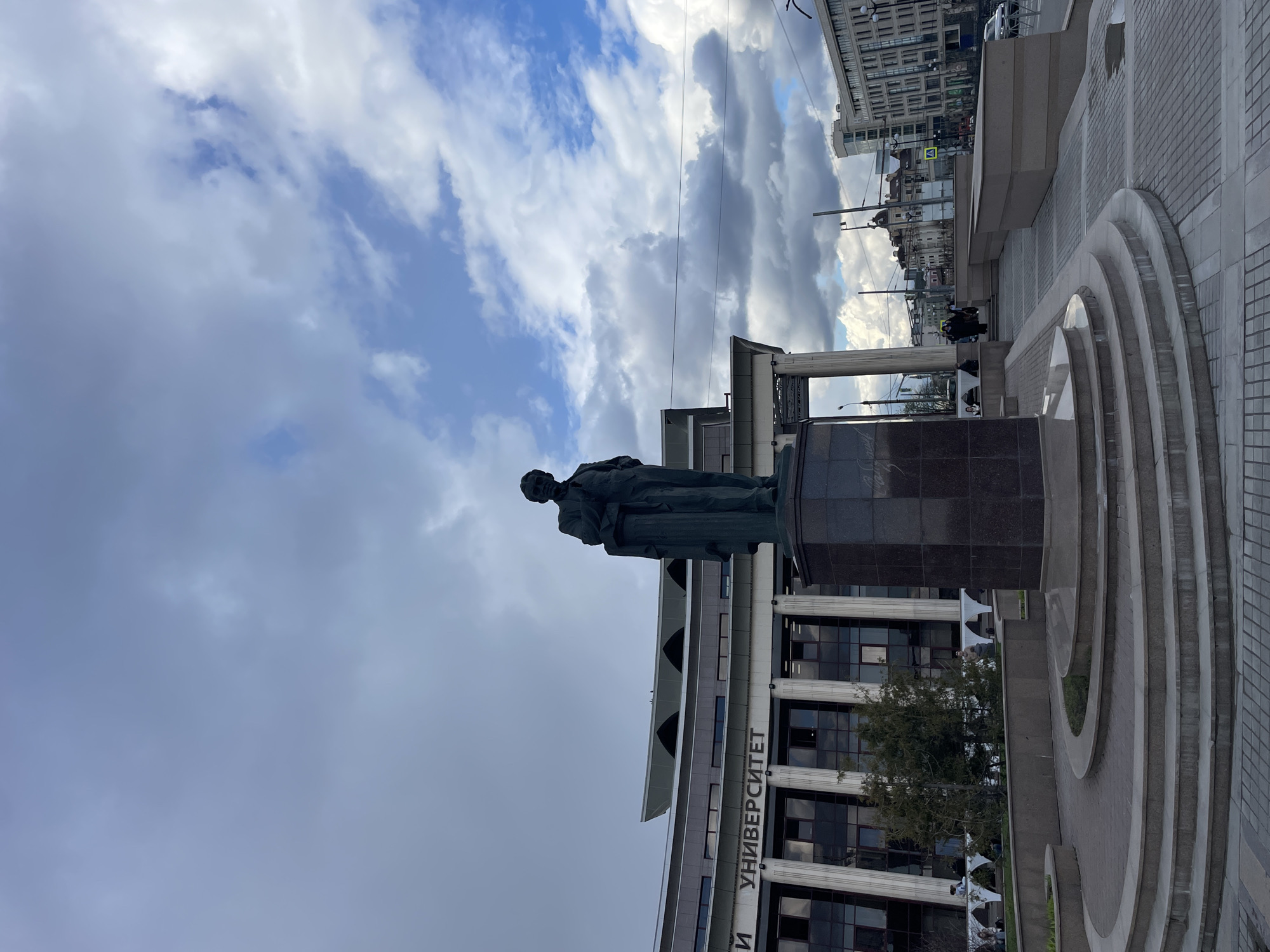 The image captures a public monument featuring a bronze statue of a man on a tall, dark rectangular pedestal. The statue depicts the man standing, dressed in what appears to be a long coat or robe. Behind the statue stands a modern multi-story building with a facade featuring large windows and white columns. The Russian word "УНИВЕРСИТЕТ" (UNIVERSITET), meaning "University," is prominently displayed on the building's exterior.
In the foreground, around the base of the monument, there are tiered, semi-circular structures made of light-colored stone or concrete, forming part of a paved plaza. A few indistinct human figures are visible in the mid-ground, walking or standing near the building and in the plaza area. Further in the background, other urban buildings and utility lines can be seen, indicating a city setting.
The sky is partly cloudy, with significant patches of both blue sky and large grey and white clouds. The lighting suggests it is daytime, likely mid-day. The scene depicts a public square or area in front of a university building, consistent with the provided context of Kazan, Russia.