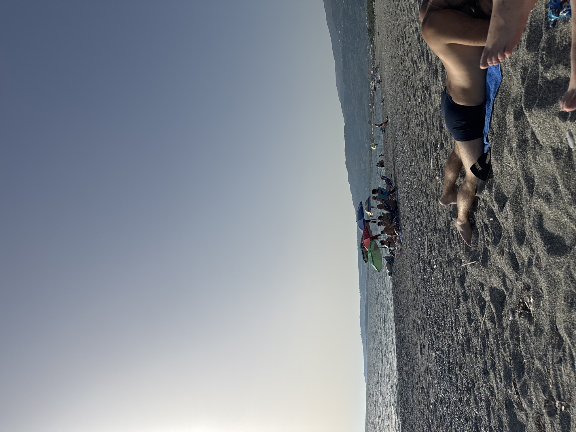 The image depicts a beach scene in Lamezia Terme, Italy, on a clear day with bright sunlight. The foreground is dominated by dark grey sand with small pebbles and some scattered debris. In the immediate foreground, a person is lying on their stomach on a blue towel, wearing dark blue swim trunks. Another person's feet are visible in the lower right corner.
Further back, along the shoreline, several beach umbrellas are set up, providing shade for groups of people. Some individuals are seated in chairs under the umbrellas, while others are lying on the sand or standing. The sea is calm with gentle waves. In the background, a hilly coastline with sparse vegetation and some buildings can be seen. The sky is clear and bright, with a hint of haze near the horizon. Several boats are visible in the distance on the water, and along the coastline.