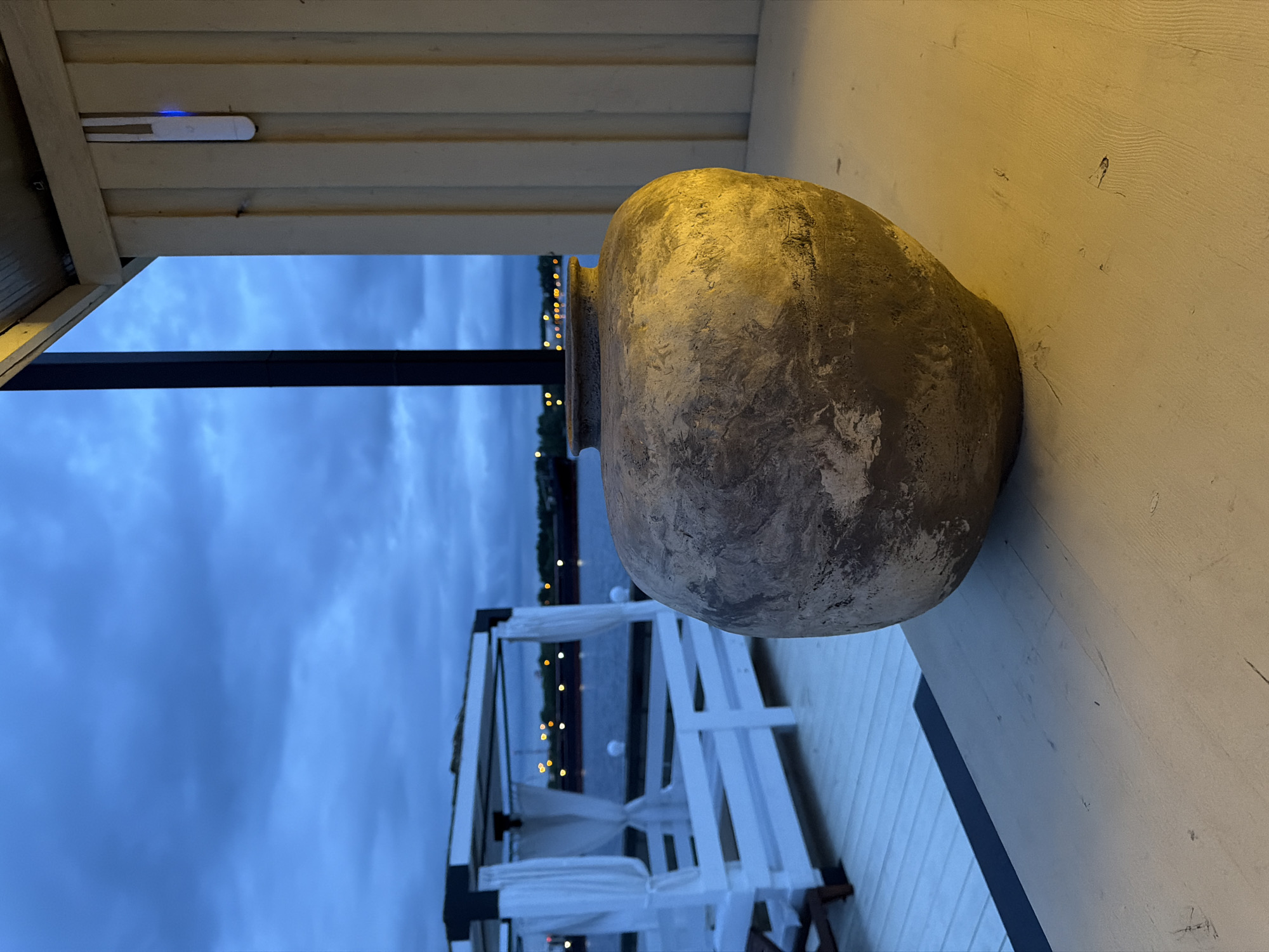 The image shows a vertical shot of an outdoor setting with a large, earthenware pot as the central focus. The pot, characterized by a rough, textured surface, sits on a weathered, white-painted wooden surface. Behind the pot, a distant view reveals a body of water, possibly a lake or river, with faint lights visible along the shoreline, suggesting a cityscape at dusk or dawn. A white gazebo-like structure with draped fabric sits near the water. The sky appears overcast, with dark blue clouds filling the upper portion of the scene. Parts of the structure with the pot are visible in the upper corner of the image.