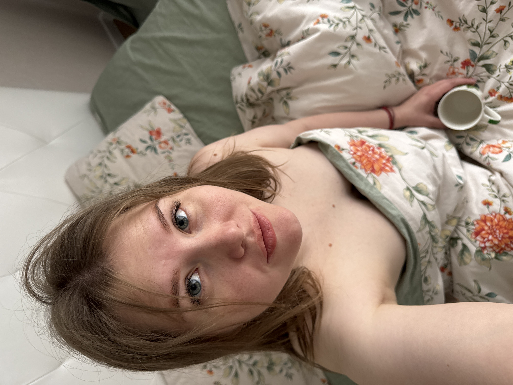 The image features a young woman lying in a bed, looking directly upwards at the camera in a selfie-style shot. She has light brown hair, fair skin, and striking blue-grey eyes, with a calm and direct expression. Her lips are slightly parted, forming a subtle, gentle smile. Her upper body, including her shoulders and upper chest, is visible, suggesting she is covered by a duvet. A few small moles are visible on her upper chest.
She is partially covered by a duvet and pillows that feature an intricate floral pattern with green vines, leaves, and prominent orange and red flowers on an off-white background. A plain light green sheet or the reverse side of the duvet is also visible. The woman holds a white ceramic mug with a green handle in her right hand, and a thin red bracelet is worn on her right wrist.
The setting appears to be indoors, likely a bedroom in Perm, Russia, during the daytime, suggested by the soft, ambient lighting. The overall scene conveys a sense of relaxation and quiet morning or leisure time. There is no visible text in the image.