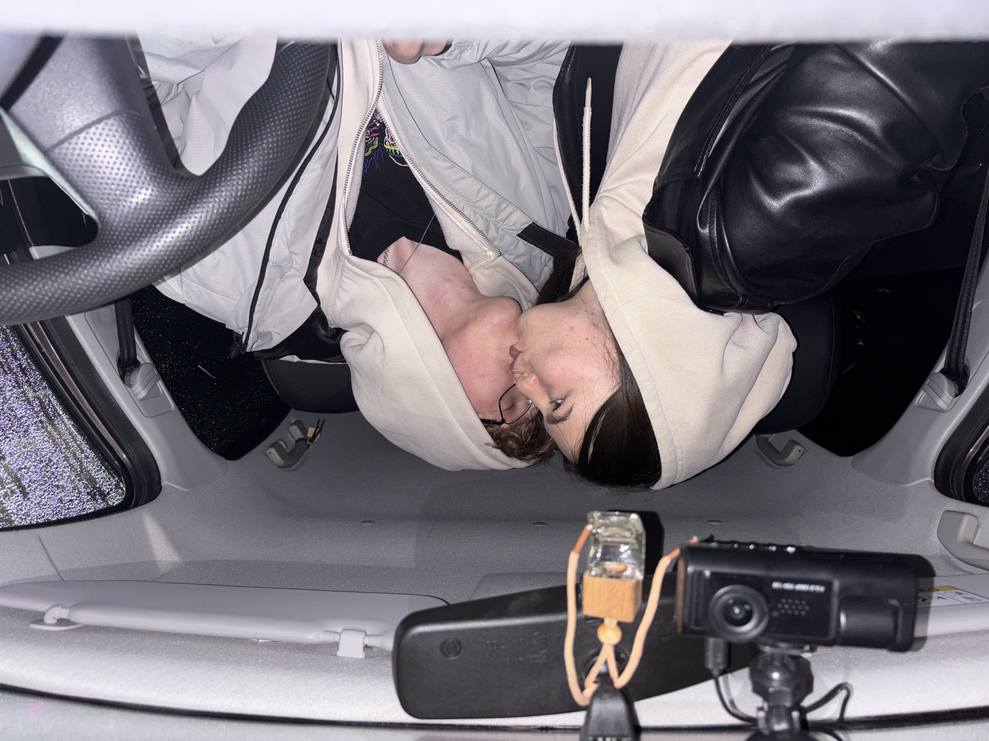 The image, taken from within a car, shows two individuals kissing. The location is identified by the presence of a dashboard camera, and a small air freshener hanging from the rearview mirror. The interior of the car shows a dark steering wheel with grey textured grip and light grey roof. On the left, a rain pattern is visible on the side window. A sun visor is in the lower left corner. The two individuals are wearing hooded jackets. One wears glasses. The location is Perm, Russia.