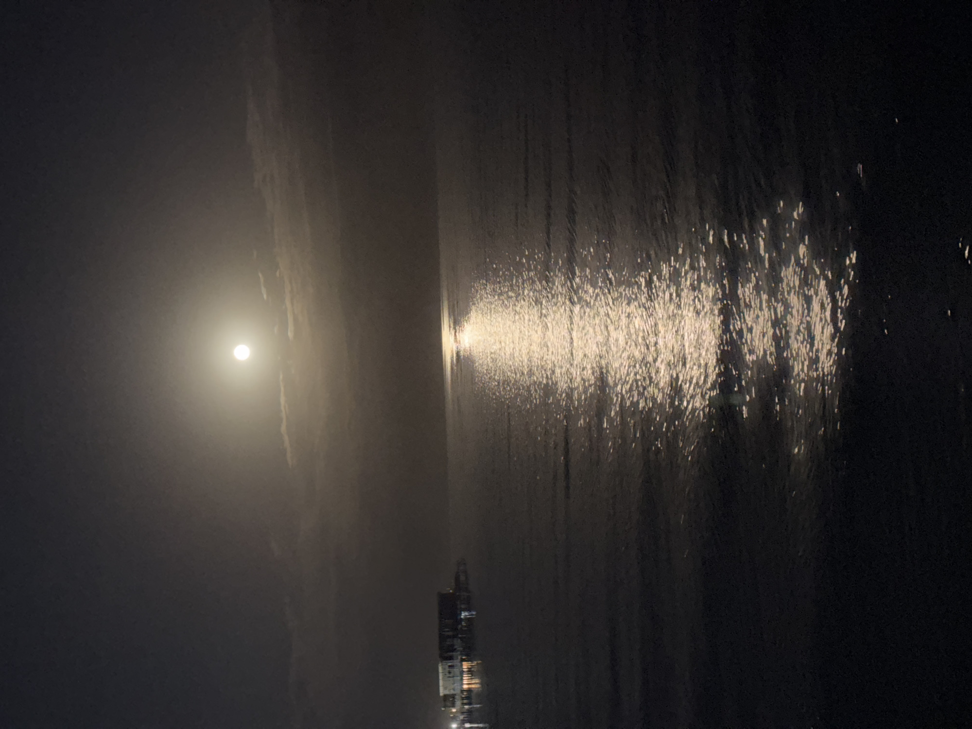 A full moon shines brightly in a dark sky, illuminating a calm sea. The moonlight creates a shimmering path across the water, leading to a distant pier or building with lights. The scene appears to be a serene night view of a coastal area, possibly in or near Sary-Oy, Kyrgyzstan.