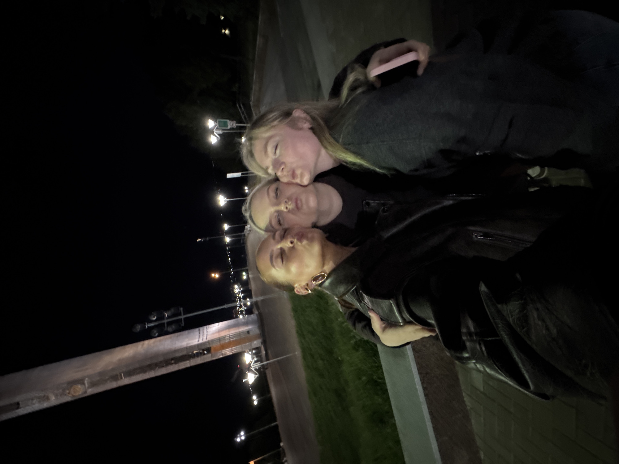 Three fair-skinned young women are posing for a photograph outdoors at night in what appears to be the city of Izhevsk, Russia.
The woman at the bottom of the frame has light blonde hair pulled back, hoop earrings, and a black leather jacket. The woman in the middle of the frame has light blonde hair and is wearing a dark top. The woman at the top has long blonde hair, a dark sweater, and is holding a phone in her right hand. All three women have their lips puckered as if giving a kiss.
In the background, streetlights are visible along a roadway and along a guardrail. There is green vegetation between the road and the guardrail.