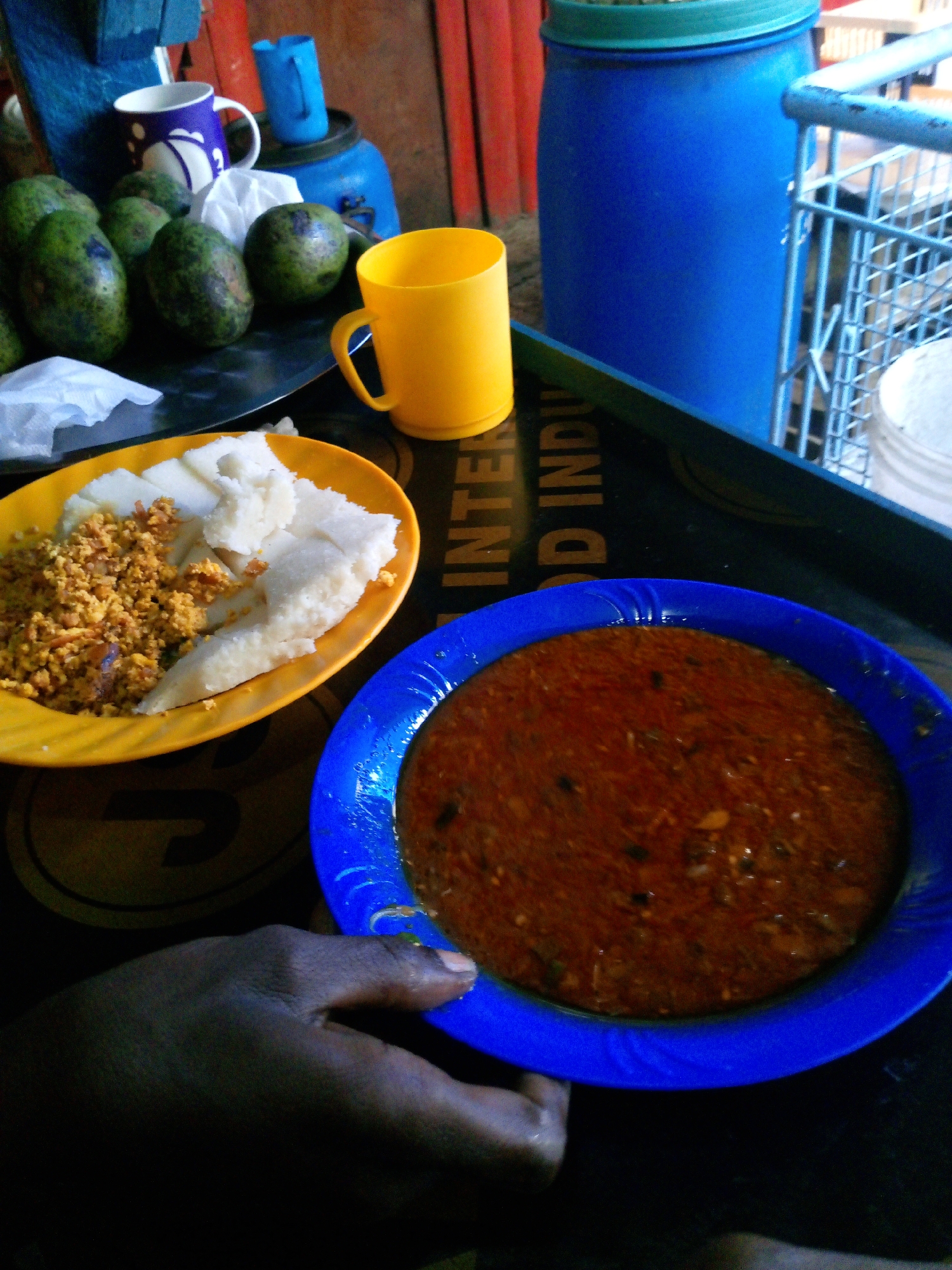 The image shows a meal on a black tray. The meal consists of a yellow plate with what appears to be mashed plantain and scrambled eggs, and a blue plate with a reddish-brown stew. A yellow mug is also on the tray, along with several avocados, and mugs, in the background. A blue barrel and a shopping cart can be seen. A hand is holding the edge of the blue plate. The location is likely a small restaurant or food stall in Nairobi, Kenya.