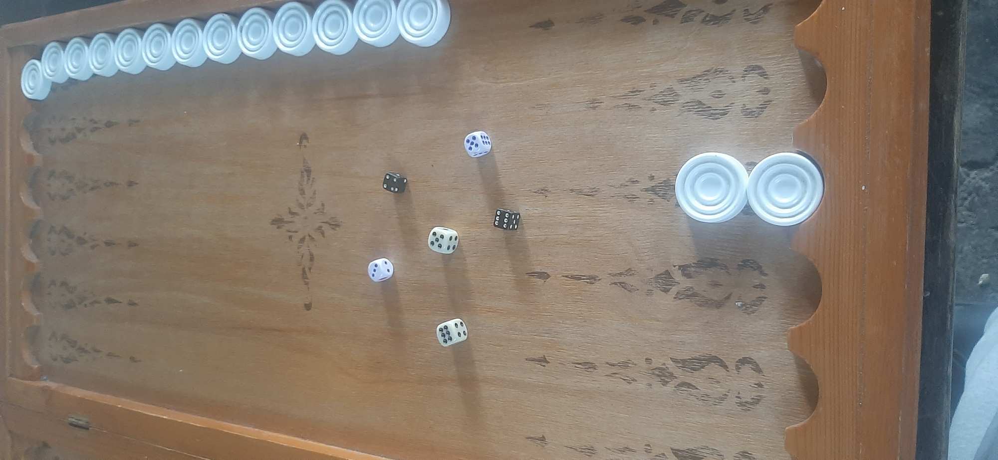 A backgammon board made of light-colored wood is displayed, with some of its playing pieces and dice scattered across the surface. Along the top edge of the board, a row of white, circular playing pieces is lined up. In the center and right portions of the board, several dice are visible. One die, with white dots on a dark background, shows the number 4. Another die, with black dots on a white background, shows the number 1. A third die, also with black dots on a white background, displays the number 3. There are also two more dice, one white with black dots showing the number 5, and another white die with black dots showing the number 3. Two white checker pieces are stacked on the right side of the board. The background of the image shows a darker, rougher surface. The scene appears to be indoors, possibly in Yessentukskaya, Russia, given the provided location context, though no specific clues to this effect are visible within the image itself. The lighting suggests it is daytime.