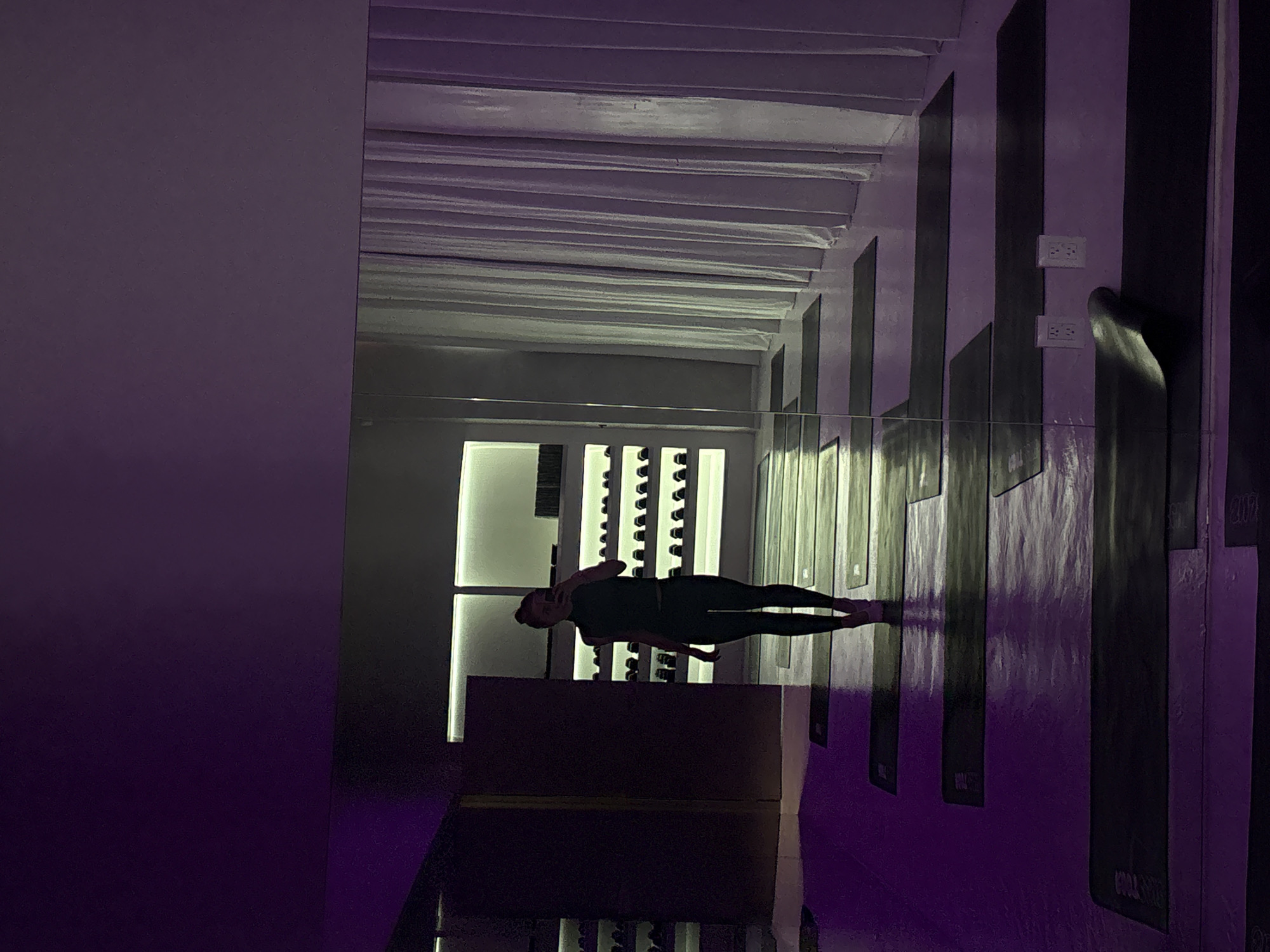 A woman is taking a full-body selfie in a dimly lit yoga studio with purple ambient lighting and white, floor-to-ceiling curtains on the right side of the frame. She is wearing dark athletic wear and her hair is pulled back. In the background, there are rows of yoga mats laid out on the floor, and shelves filled with what appear to be water bottles. The studio has a modern, minimalist aesthetic with a large mirror dominating the left side of the frame, reflecting the woman and the studio space. The location is identified as Mexico City, Mexico.