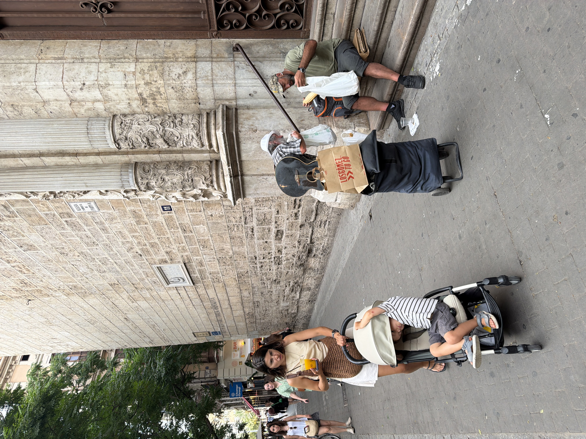 An eye-level, medium shot captures a street scene with a woman pushing a stroller in the foreground, walking past a man sitting on steps with a guitar and shopping bags. The woman, wearing a beige sleeveless top and white shorts, holds a cup of orange juice in her right hand and pushes a beige stroller with a child inside. The child, wearing a striped shirt and gray shorts, reclines in the stroller. The man sitting on the stone steps wears a green t-shirt, gray shorts, a camouflage baseball cap, and black sneakers. He is holding a white plastic bag with bread. Beside him are a large black guitar with a brown neck, a shopping cart with several bags, and a small plastic cup on the ground. The background features a textured stone building with ornate columns and a large wooden door. Several other people are visible in the distance, walking along the cobblestone street lined with trees and buildings. The lighting suggests it is daytime, likely overcast or late afternoon.