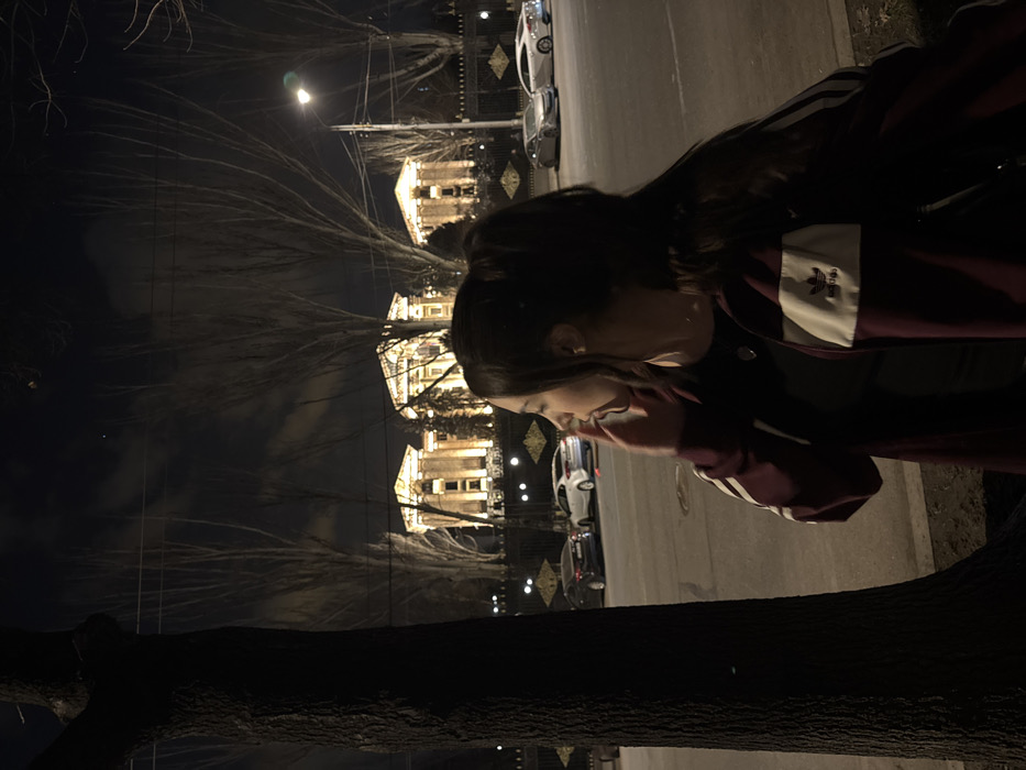 A woman is photographed outdoors at night. She wears an Adidas jacket, and her hair is up. In the background are three lighted buildings with decorative fencing and streetlights, plus parked cars on the right side of the frame. A dark tree trunk obscures the lower left corner. The image has been rotated.