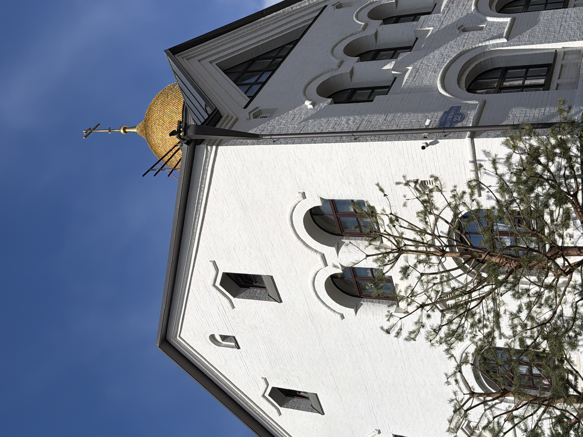 The image captures a building in Moscow, Russia. The building is white and features rounded windows and triangular accents. A golden dome with a cross tops the structure, indicating a religious affiliation, possibly a church. A tree with green needles partially obscures the view of the building. A blue street sign is visible near the top of the building. The sky is a clear blue.