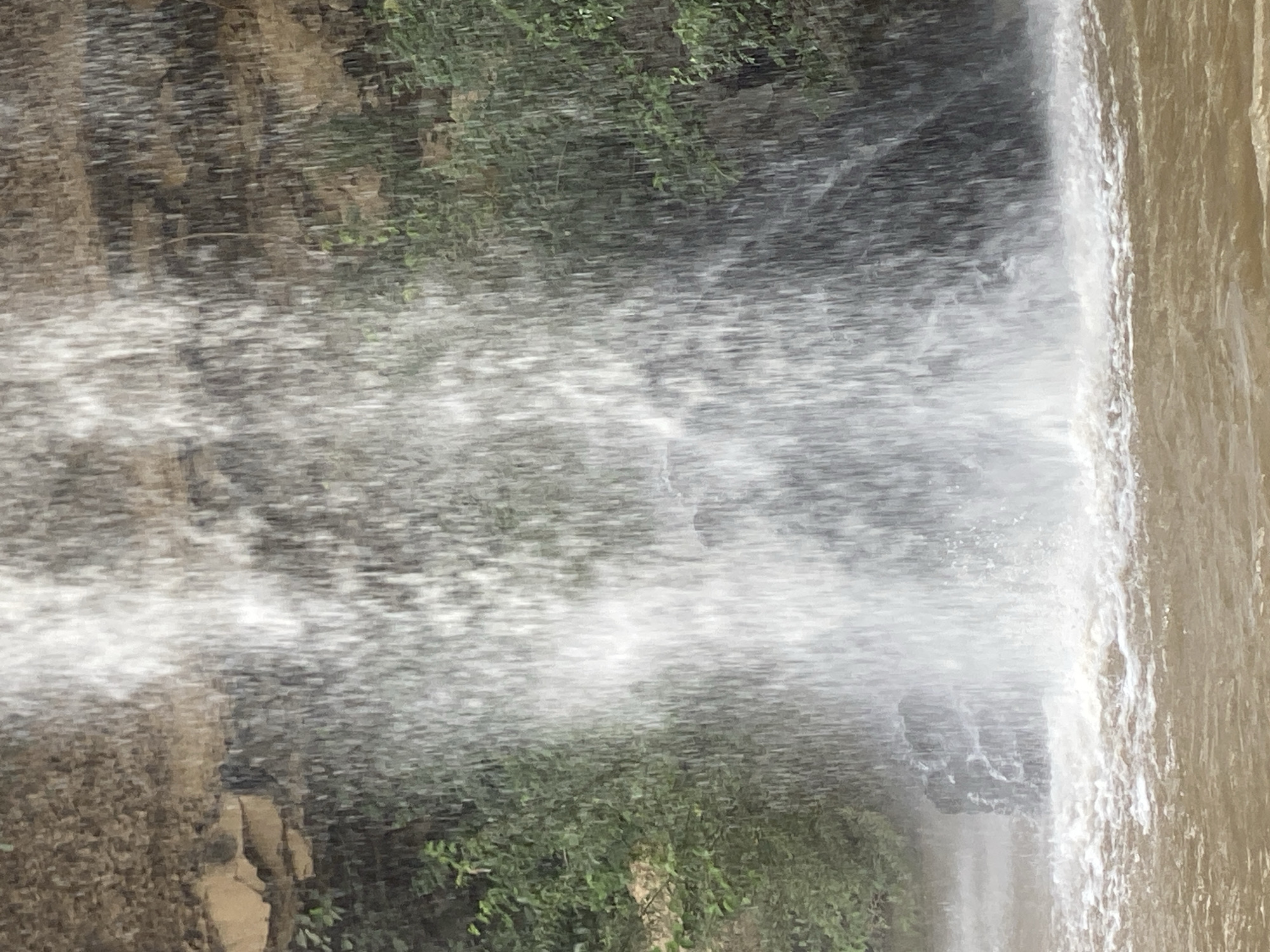 A vertically oriented image captures a powerful waterfall in a natural, outdoor setting, likely near Maigizo, Nigeria. The scene is dominated by a large volume of cascading water. On the left and central portions of the frame, the water appears as a dense, white curtain of spray and mist, indicating a strong downward flow. To the far right, a distinct section of the main water body is visible, characterized by its muddy brown color, which sharply contrasts with the white spray.
Behind the curtain of water, partially obscured by the mist, green foliage and brown, rugged textures of rocks or earth can be discerned, suggesting a vegetated cliff or riverbank. The bright lighting indicates it is daytime. No people, specific man-made objects, or visible text are present in the image. The focus is entirely on the dynamic movement and force of the natural water feature.