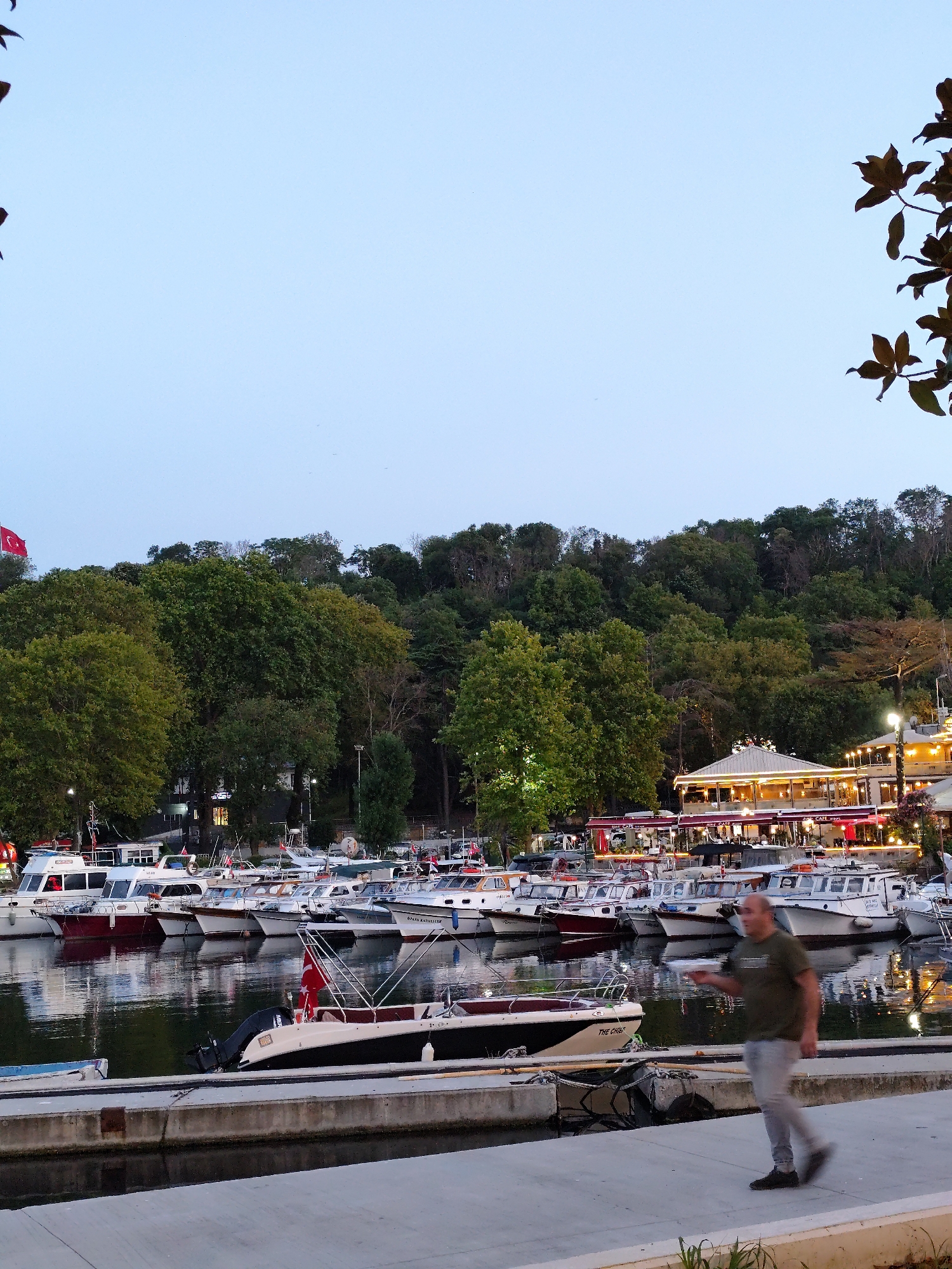 The image depicts a waterfront scene in Sarıyer, Turkey, at dusk. In the foreground, a concrete promenade lines a calm body of water where numerous boats are docked. A man in a green t-shirt and grey pants is walking on the promenade from right to left, appearing to hold a white object in his right hand. Behind the boats, a lush, tree-covered hillside rises, punctuated by buildings and lights. One prominent building features a white roof and is illuminated with string lights, suggesting it is a restaurant or cafe. A Turkish flag flies on a pole to the left of the frame, further identifying the location. The water reflects the lights from the buildings and the sky is a soft blue, indicating the end of the day.
