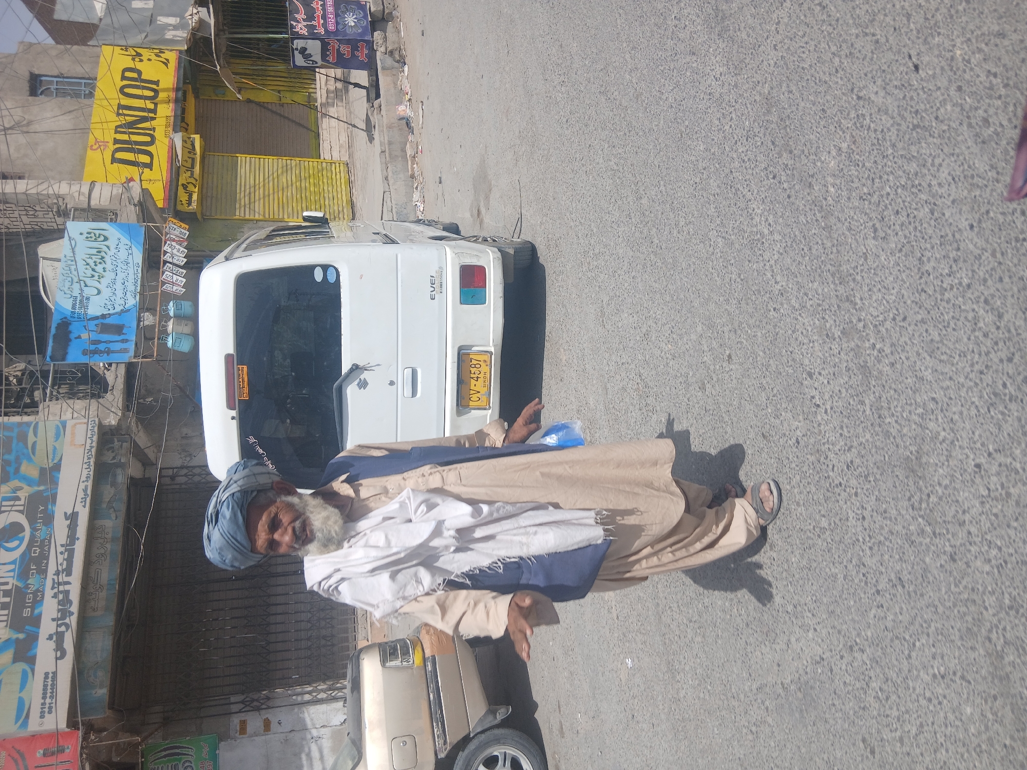 A street scene in Quetta City Tehsil, Pakistan. An elderly man with a white beard and wearing a blue turban, a light brown robe, and sandals, stands on the street near the rear of a white van with license plate CV-4587. Various storefront signs with product offerings in both English and Urdu are visible, including "DUNLOP" and auto parts signage. The buildings appear weathered, and the street is asphalt. The camera angle seems rotated.
