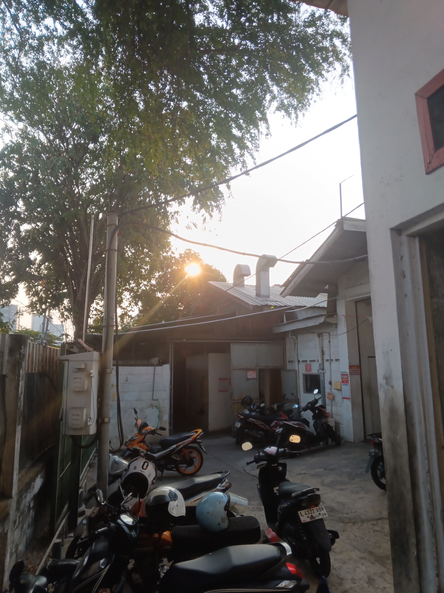 The image depicts an outdoor scene, likely in Surabaya, Indonesia, during a late afternoon or early evening with the sun setting behind a large tree and buildings. The foreground is dominated by several parked motorcycles, with their handlebars, seats, and some helmets visible. One motorcycle in the center has a license plate that reads "L 4322 ABR 06-28".
Behind the motorcycles, a single-story building with a corrugated metal roof is visible. Various pipes and ventilation systems are present on the roof. The building's exterior is painted white, and there are several signs and notices posted on the wall. Adjacent to this building, another structure, possibly a wall or another part of the building, is made of concrete blocks.
A tall, slender utility pole stands prominently to the left of the center, with wires running from it. The large tree, with its dense foliage, occupies the upper left portion of the image, casting shadows and allowing sunlight to filter through its leaves. The sky is bright, indicating the sun is low on the horizon. The overall environment suggests a commercial or industrial area, possibly a back alley or a parking lot associated with businesses. No people are visible in the image.