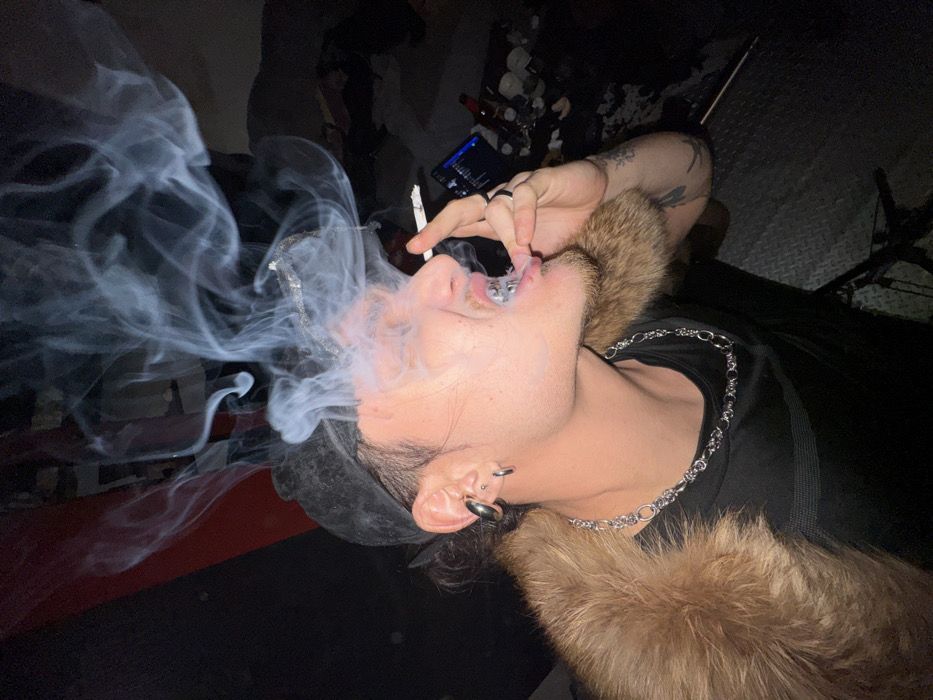 A young adult male is depicted in a profile shot, facing left and exhaling a dense plume of smoke from his mouth. He wears a dark baseball cap backward, a dark t-shirt, and a prominent brown fur collar draped over his shoulders. A silver-colored link chain necklace is visible around his neck. His right ear features a large silver hoop or gauge in the lobe, a smaller silver hoop in the helix, and a stud in the upper cartilage. He has a short beard and is sporting a silver-colored grill on his lower teeth. His right hand, adorned with rings, holds a lit cigarette between the thumb and forefinger, positioned near his lower lip. Indistinct tattoos are visible on his right forearm. The background is largely dark and underexposed. To the immediate left, a dark red or maroon vertical surface is present. To the right and rear of the subject, a cluttered area reveals a screen displaying a blue-lit interface, possibly a music application, along with a dark bottle and various other unidentifiable items on a dark surface. The floor in the lower right appears dark and textured. The specific location within the city of Sofia, Bulgaria, cannot be identified from the visual information provided.