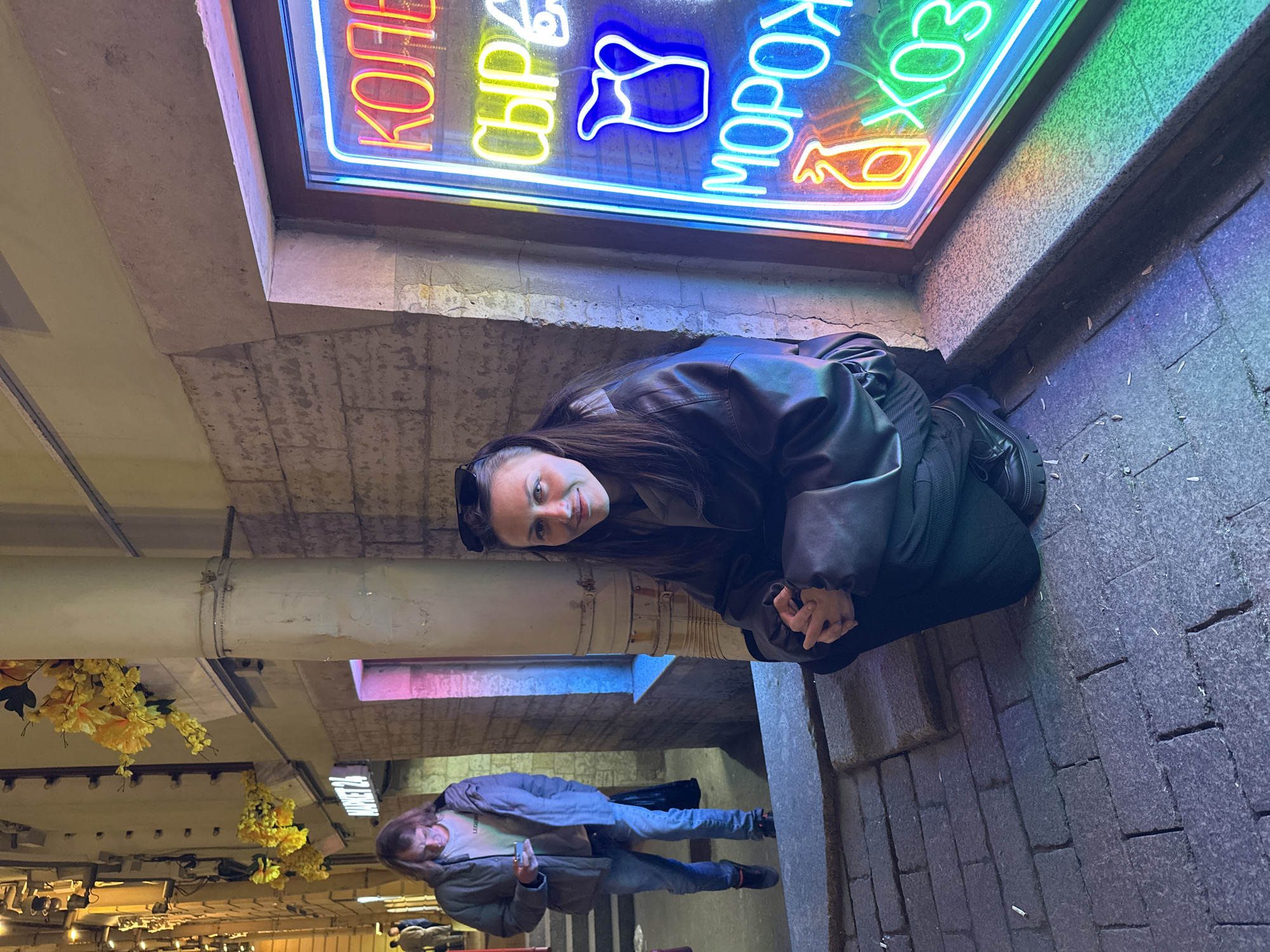 A woman in a black leather jacket is kneeling next to a building's foundation in Saint Petersburg, Russia. Above her, a neon sign reads "КОФЕ СЫР МОРОЖЕНОЕ ХОЗ." A man, dressed in a light jacket and jeans, stands in the background, looking at his phone. Yellow flowers hang from the ceiling. The pavement is visible beneath the woman.